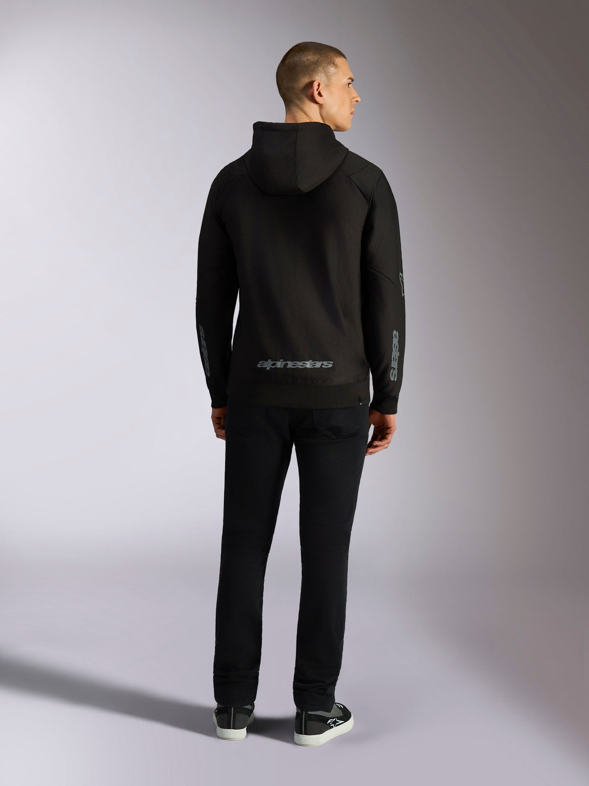 Alpinestars Sessions V3 Hoodie, vardaglig streetwear, svart med grått Alpinestars-ordmärke på nedre delen av ryggen och 'astars'-logotyp på ärmarna, ryggvy av manlig modell som bär huvtröja och svarta byxor, modern motorsport-inspirerad design