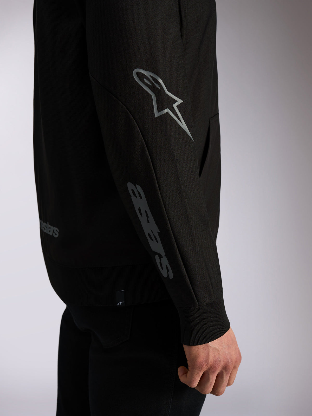 Alpinestars Sessions V3 Hoodie, svart ledig streetwear, närbild på ärmen med grå Astars-logotyp på armbågen och textlogotyp på underarmen, ribbade muddar, motorsport-inspirerad design