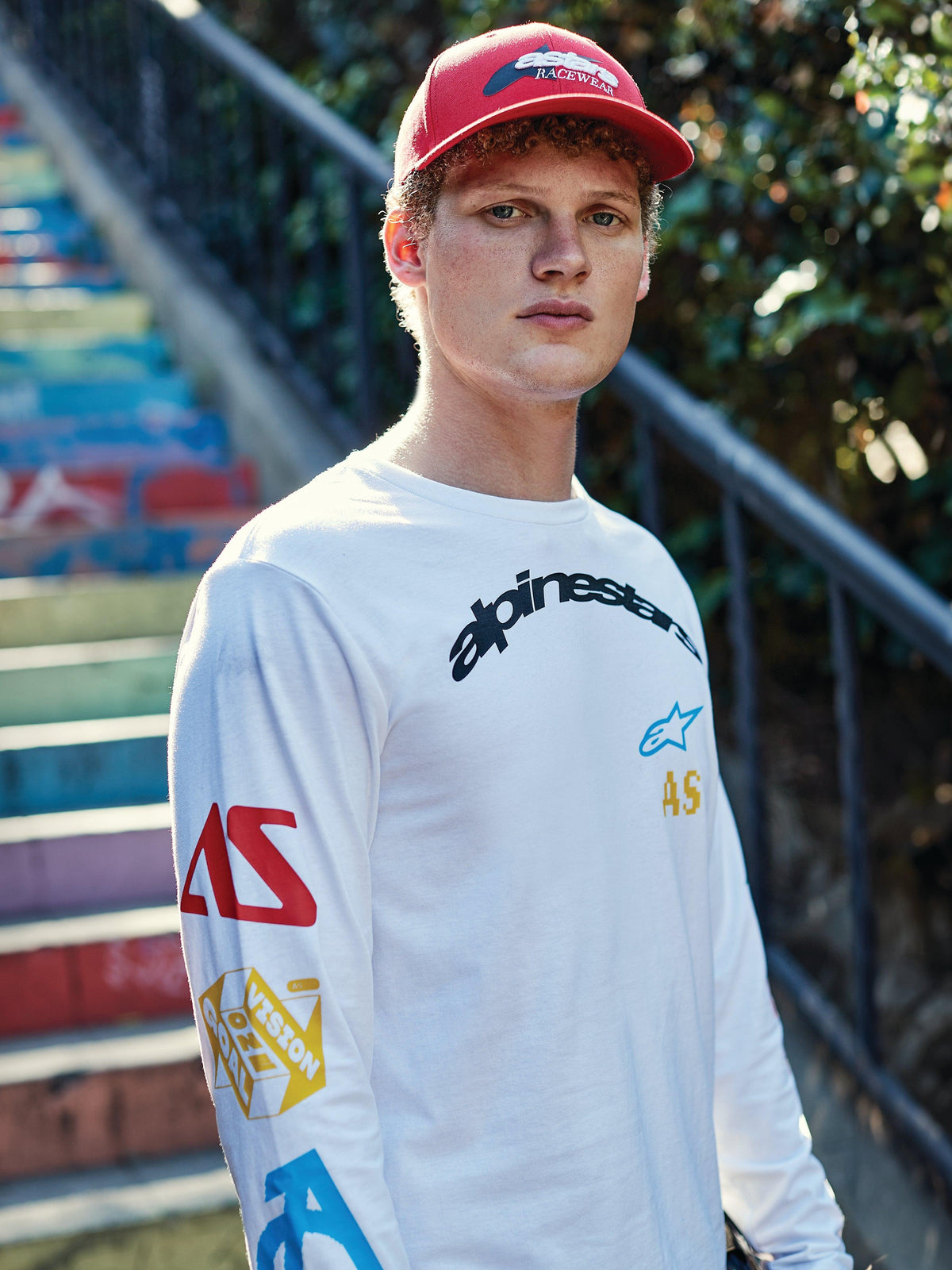 Alpinestars Profile keps, röd, bärs av en ung man, strukturerad krona med böjd skärm och broderad logotyp, matchad med en vit långärmad t-shirt med grafiskt tryck, avslappnad motorsport-streetwear
