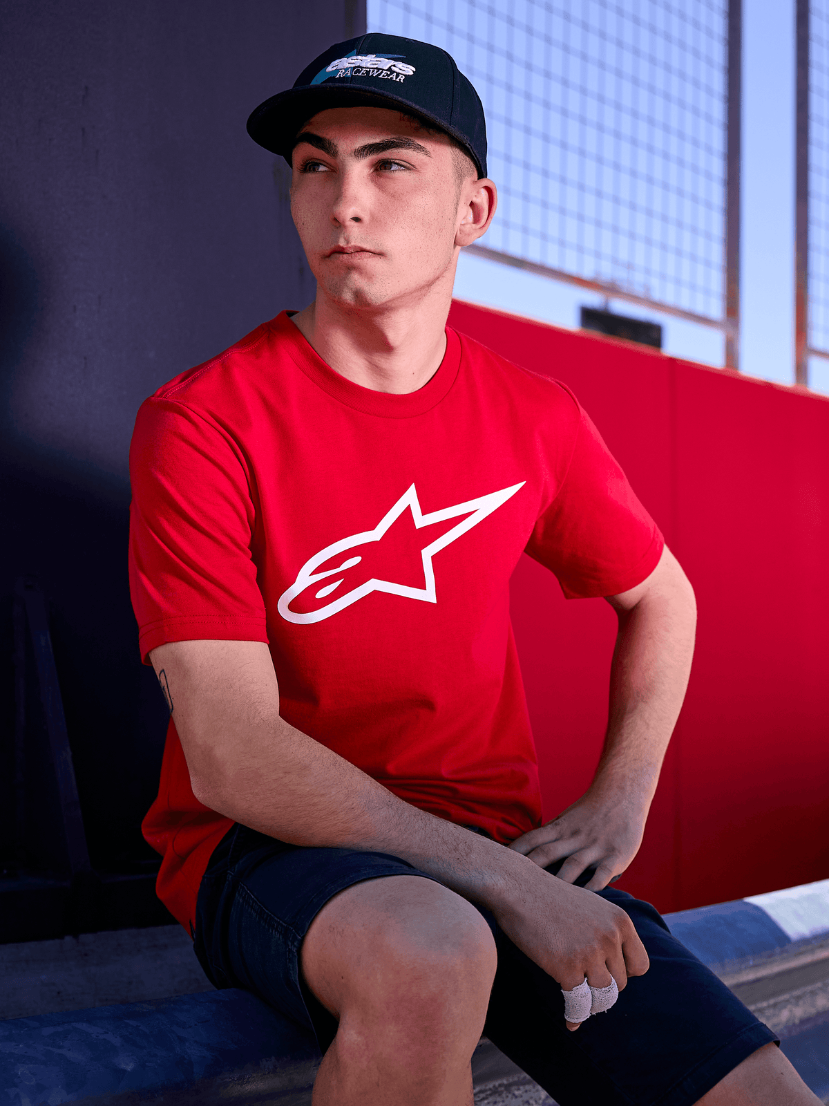 Alpinestars Profile Hat i svart, livsstilsbild av en man som bär kepsen med böjd skärm och en röd t-shirt med vit Alpinestars-logotyp, vardagligt motorsportinspirerat streetwear