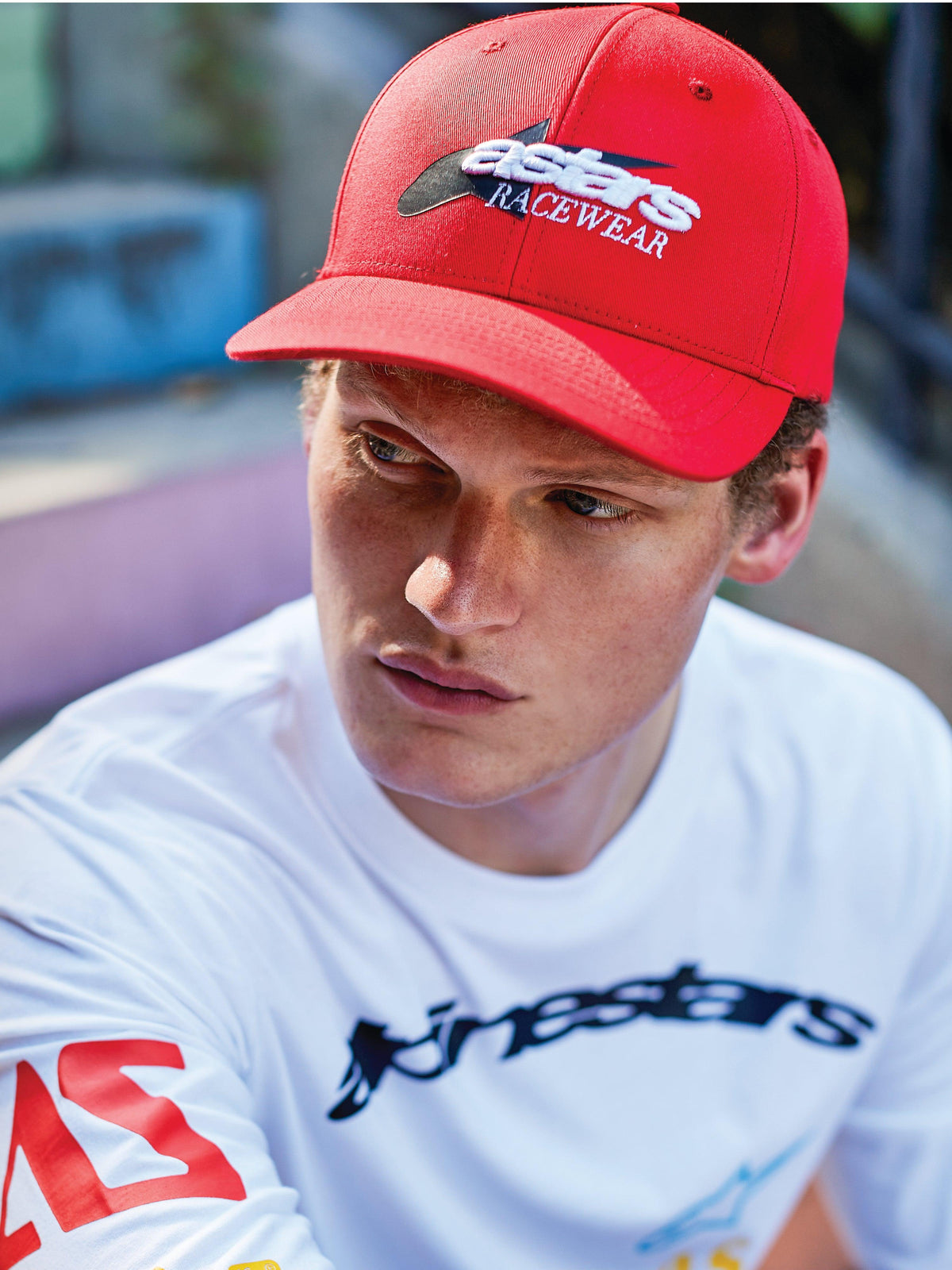 Alpinestars Profile Hat, röd snapback-keps med böjd skärm, med vit och svart 'astars RACEWEAR'-brodyr fram, kombinerad med en vit långärmad Alpinestars-tröja med grafik, avslappnad motorsport-livsstil