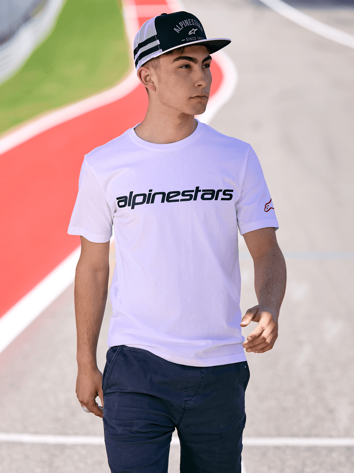 Man som bär Alpinestars Backline Trucker Hat i svart och vitt, kombinerad med en vit Alpinestars t-shirt med logotyp och mörkblå byxor, avslappnad motorsport-streetwear på en utomhusbana