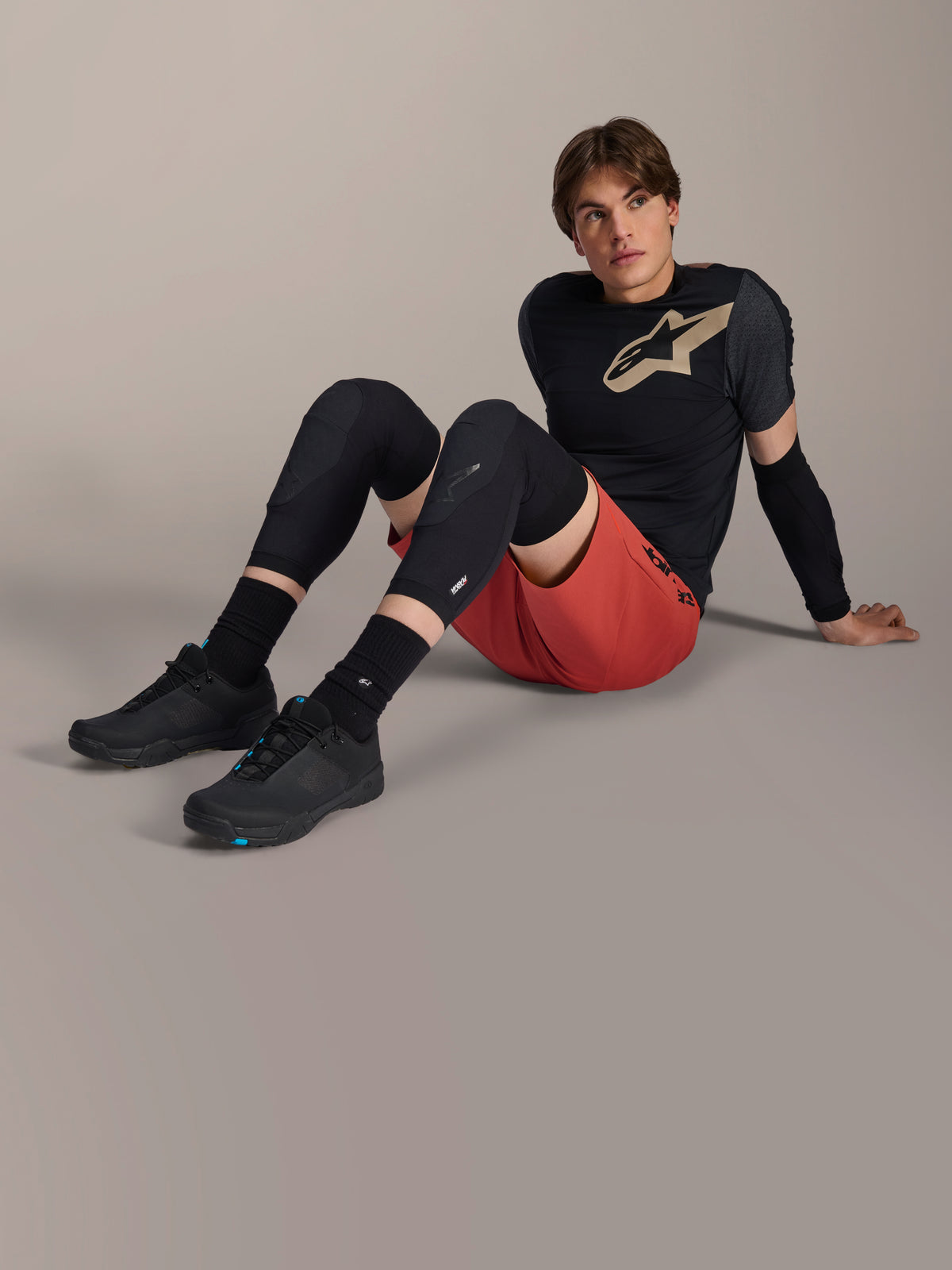 Alpinestars A-Motion Plasma armbågsskydd, bärs av en manlig cyklist som sitter på ett studiogolv, med svart mesh-konstruktion, kombinerat med röda shorts och en svart Alpinestars-tröja med beige logotyp, högpresterande mountainbike-skydd för trailcykling
