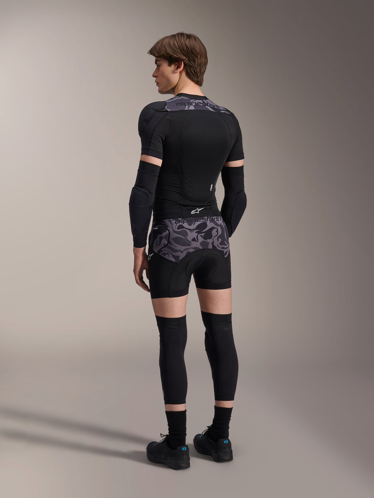 Modell som bär Alpinestars A-Motion Plasma skyddsboxershorts i Black Camo och matchande A-Motion Plasma skyddsjacka, vy bakifrån som visar grått kamouflagemönster, armbågsskydd, knäskydd och cykelskor, komplett uppsättning skyddsutrustning för mountainbike.