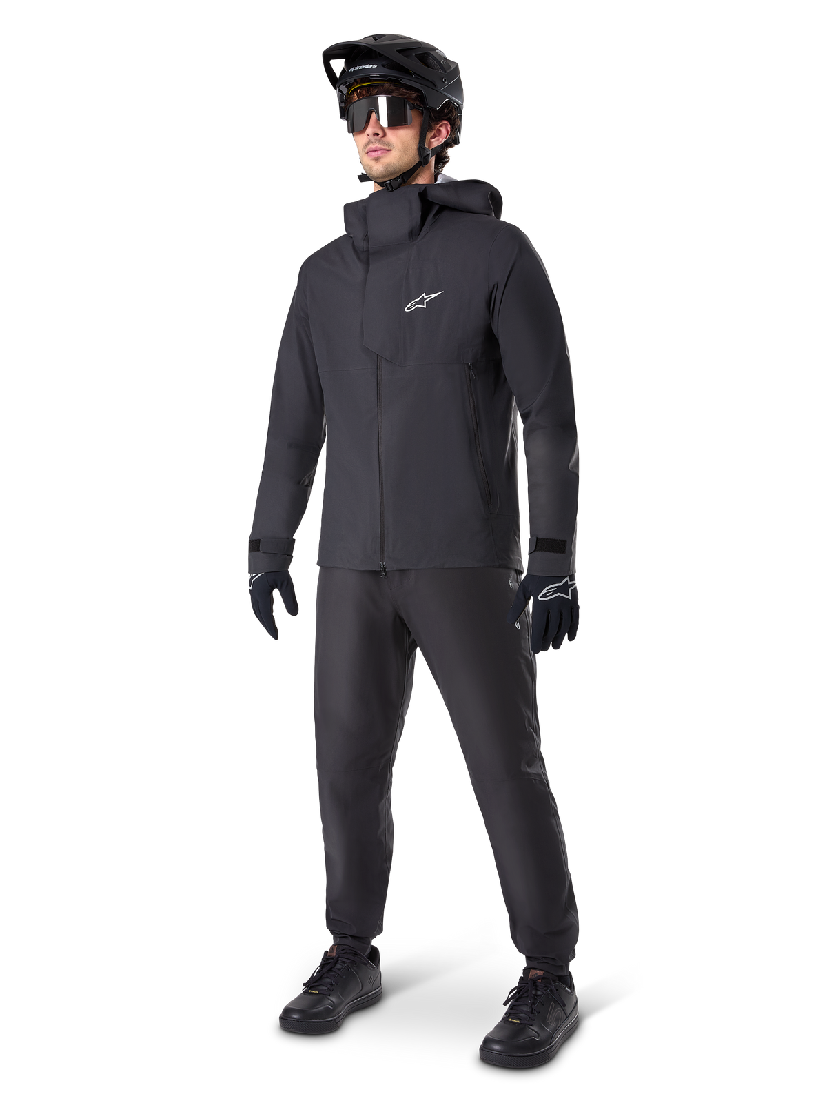 Manlig mountainbike-cyklist klädd i svarta Alpinestars A-Dura Waterproof Pants, matchande svart vattentät jacka med huva, svart MTB-hjälm med visir, mörka cykelglasögon och svarta handskar, komplett professionellt utrustningsset för terrängcykling i alla väder