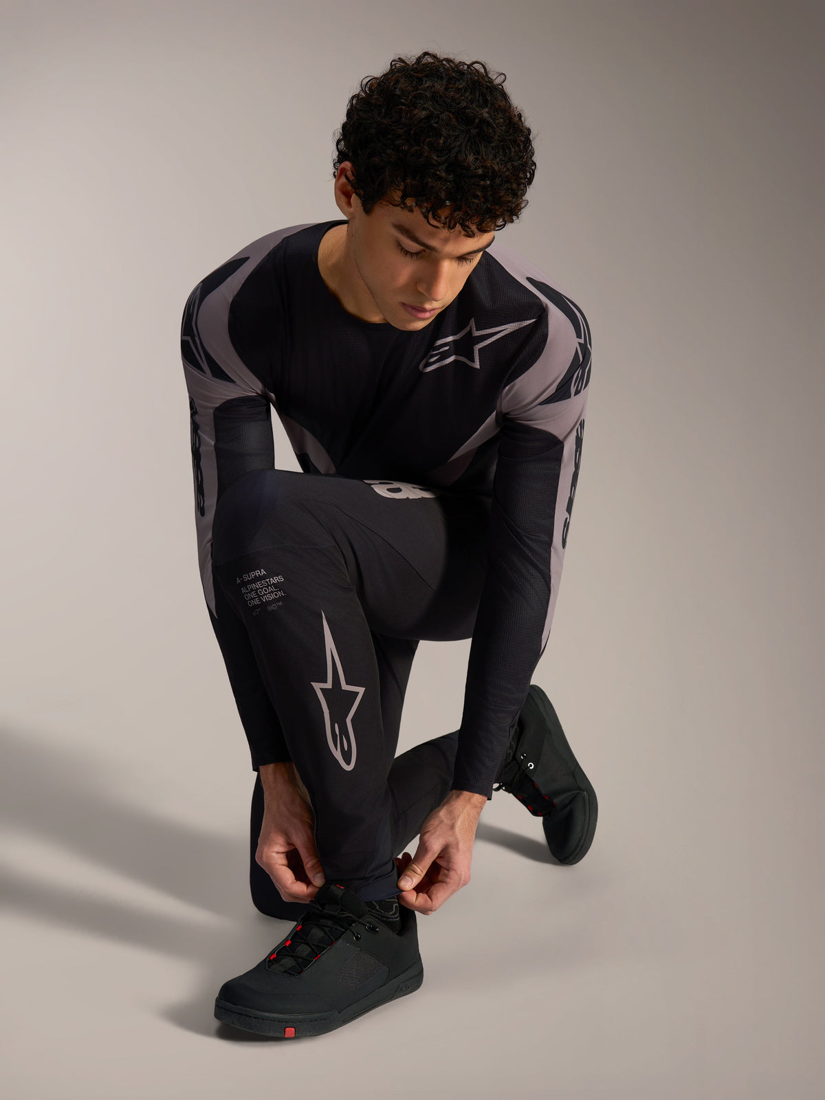 Svarta Alpinestars A-Supra Elite Byxor med grå racinglogotyper på en knäböjande modell för MTB-cykling.