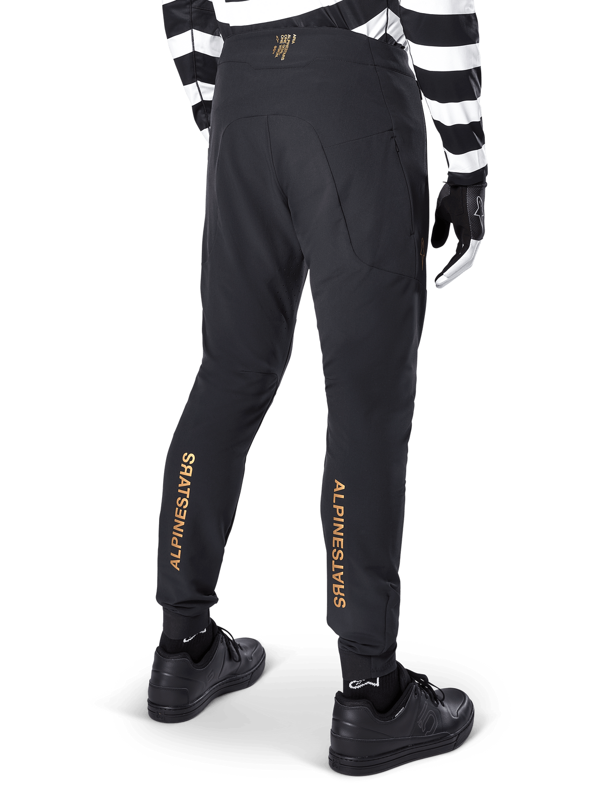 Alpinestars A-Aria Elite byxor, svarta MTB-byxor, vy bakifrån, med vertikal Alpinestars-logotyp i guld på baksidan av benen, bärs med en svart- och vitrandig tröja och svarta skor, högpresterande cykelkläder för mountainbike