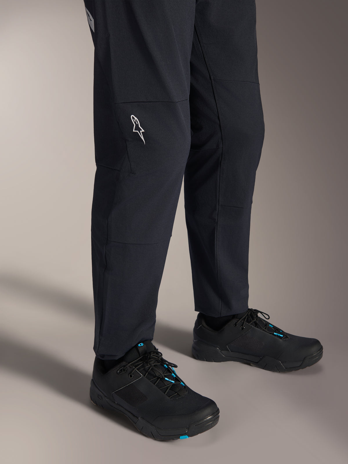 Alpinestars A-Dura Elite-byxor, svarta mountainbikebyxor, vy av underbenen med en vit Alpinestars-logotyp på knät, bärs med svarta och blå cykelskor, slitstark konstruktion för MTB-cykling