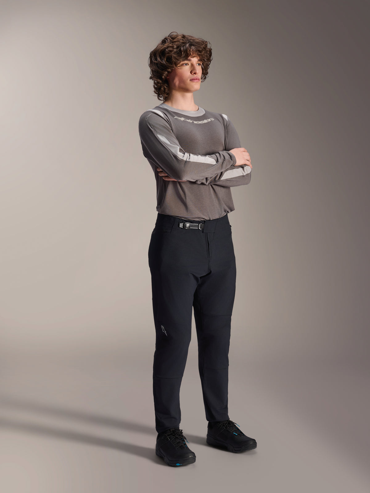 Manlig modell i Alpinestars A-Dura Elite-byxor, svarta MTB-byxor, matchade med en grå långärmad tröja och svarta cykelskor, spärrspänne framtill, professionella mountainbikekläder för trail- och endurocykling