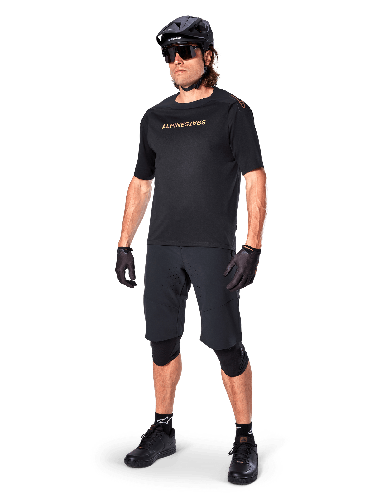 Cyklist som bär Alpinestars A-Aria Elite Shorts, svarta mountainbikeshorts, matchat med en svart tröja med logotyper i guld, svart hjälm, solglasögon och knäskydd, komplett outfit för terrängcykling