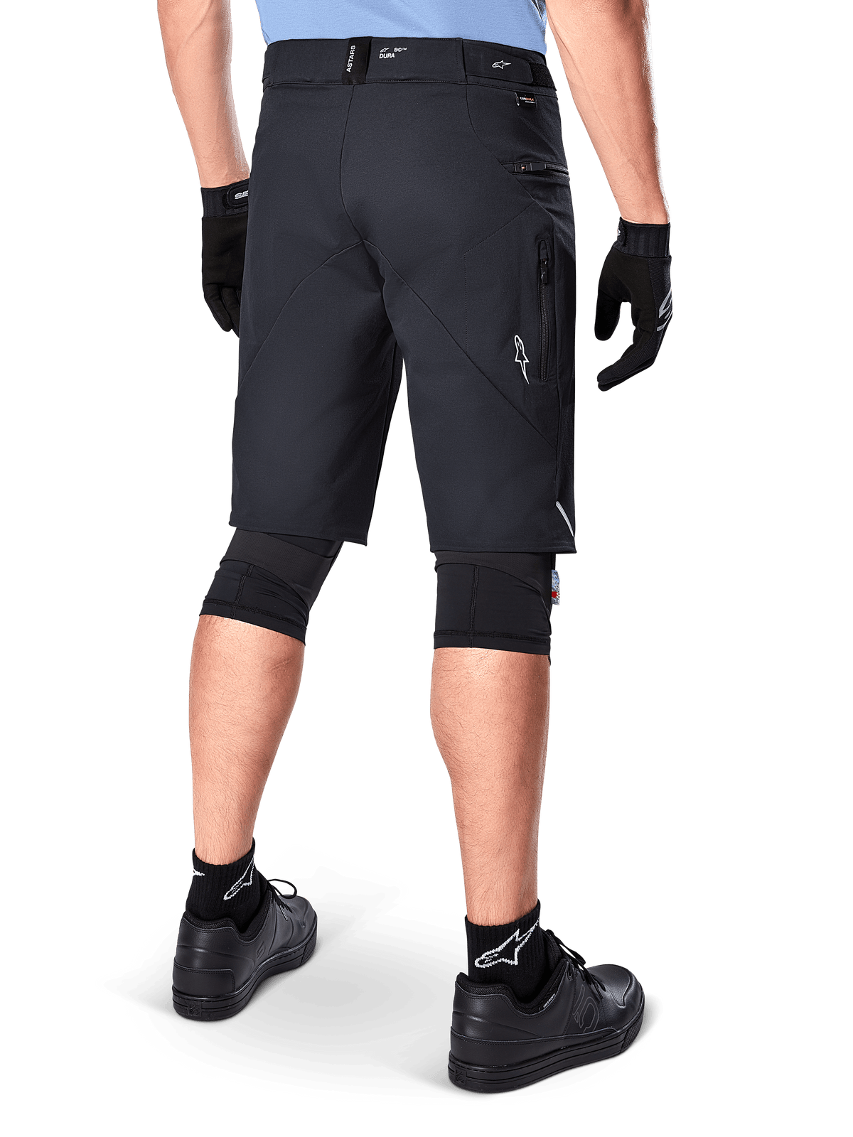 Alpinestars A-Dura Elite Shorts, Black, rear view of a rider wearing mountain bike gear including black knee pads, gloves, and socks with white logos, featuring zippered side pockets and reflective accents on a durable Cordura fabric.
