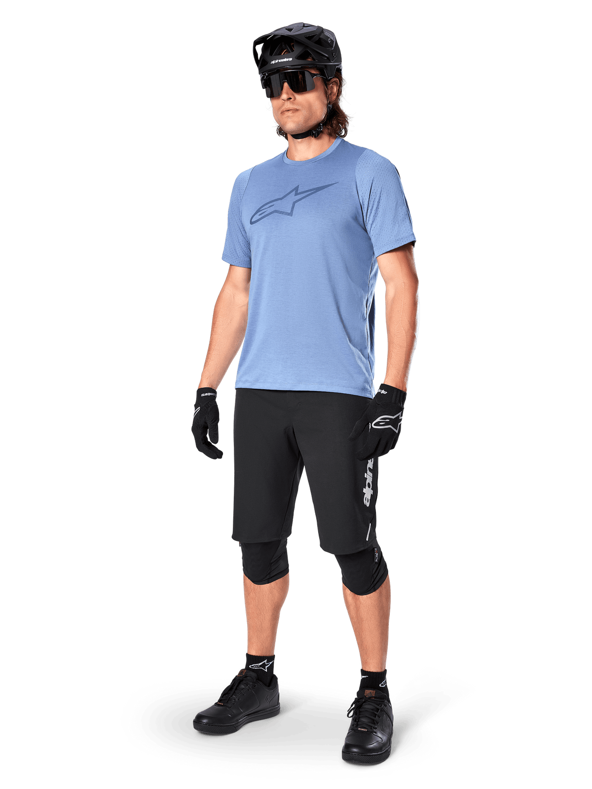 Male rider wearing Alpinestars A-Dura Elite Shorts in Black, light blue short-sleeve jersey with chest logo, black helmet, sunglasses, and protective knee pads, full mountain biking gear setup for off-road trails.