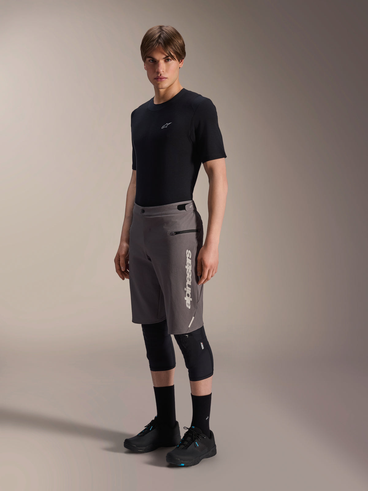 Alpinestars A-Dura Elite Shorts, mörkgrå mountainbikeshorts, bärs av en modell med en svart t-shirt och knäskydd, med en vertikal vit Alpinestars-logotyp på vänster ben, tekniskt Cordura-tyg för terrängcykling