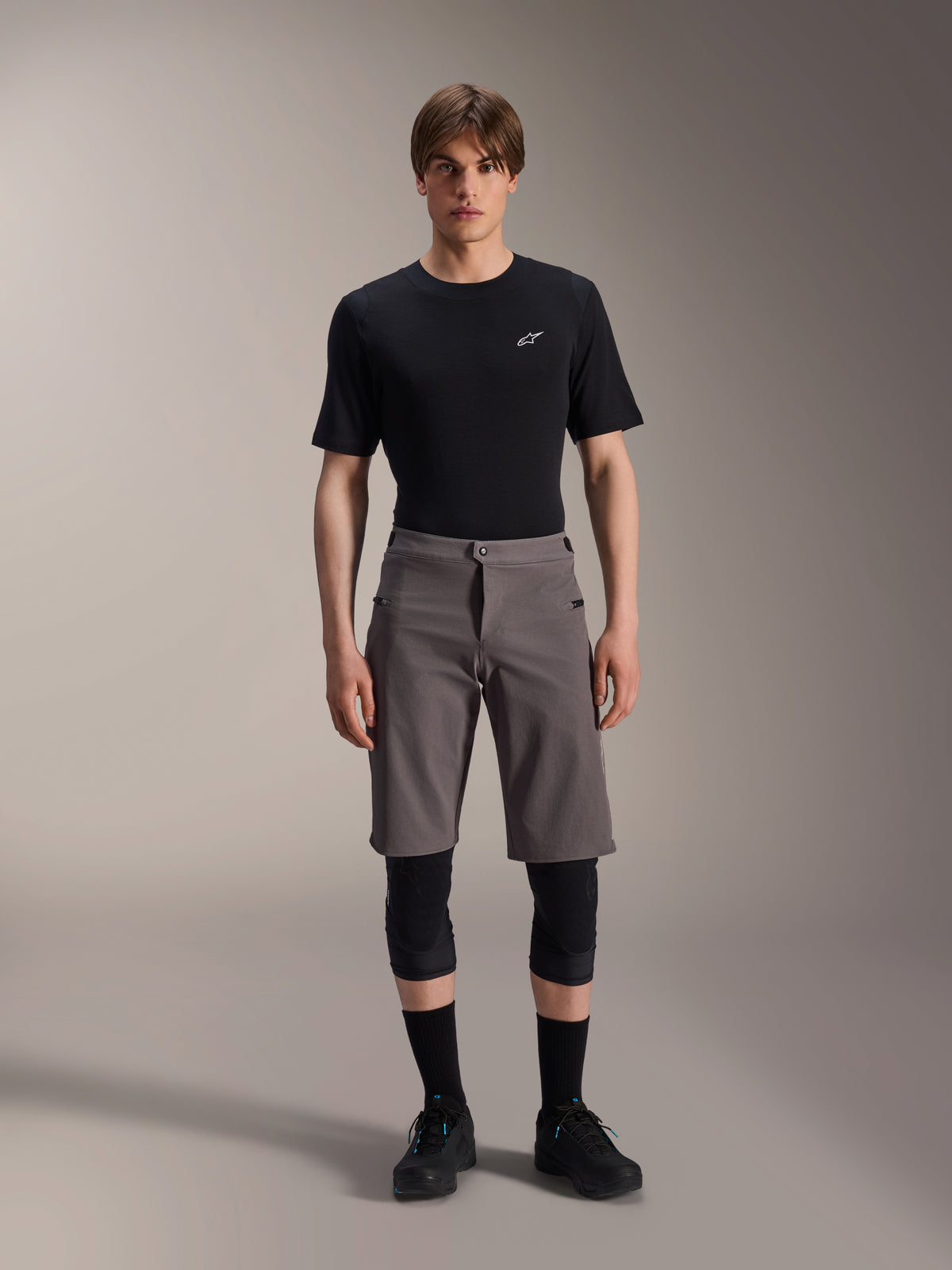 Alpinestars A-Dura Elite Shorts, mörkgrå MTB-shorts, bärs med en svart t-shirt med rund halsringning och svarta knäskydd, knälånga funktionskläder för cykling med tryckknappsstängning fram och slitstarkt Cordura-tyg.