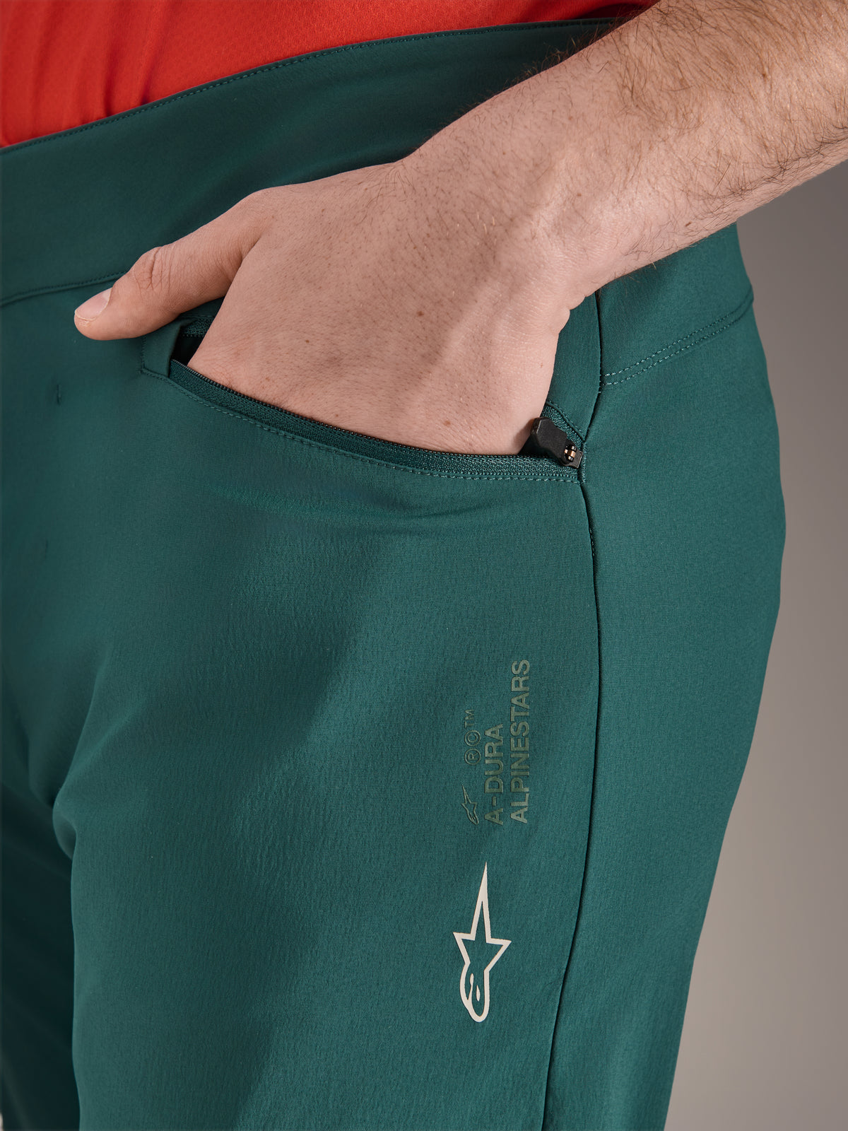 Alpinestars A-Dura byxor i färgen Dark Pine, närbild på blixtlåsförsedd ficka med hand, med vit Alpinestars-logotyp och text på låret, slitstarka MTB-cykelbyxor