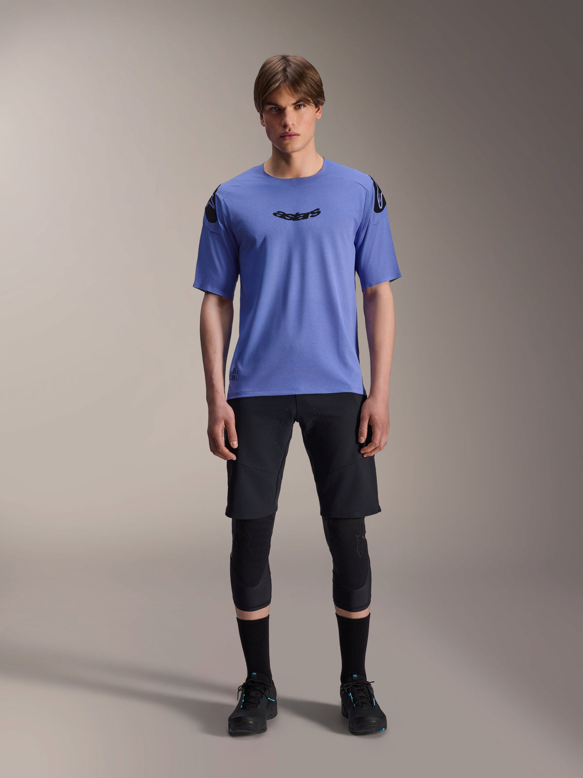 Male model wearing Alpinestars A-Aria Polartec Elite Short Sleeve Jersey in Baja Blue, black astars logo on the chest, paired with black MTB shorts, knee guards, and black cycling shoes, studio shot for mountain bike apparel