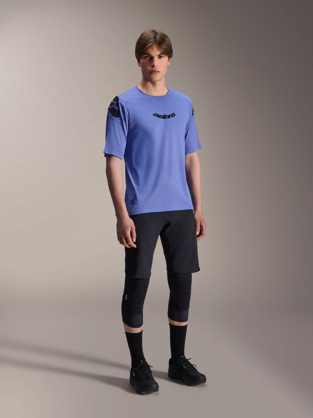 Male model wearing Alpinestars A-Aria Lite Shorts in black, paired with a blue A-Aria Polartec Elite Short Sleeve Jersey, black knee guards, and black cycling shoes, performance mountain bike apparel for trail riding.