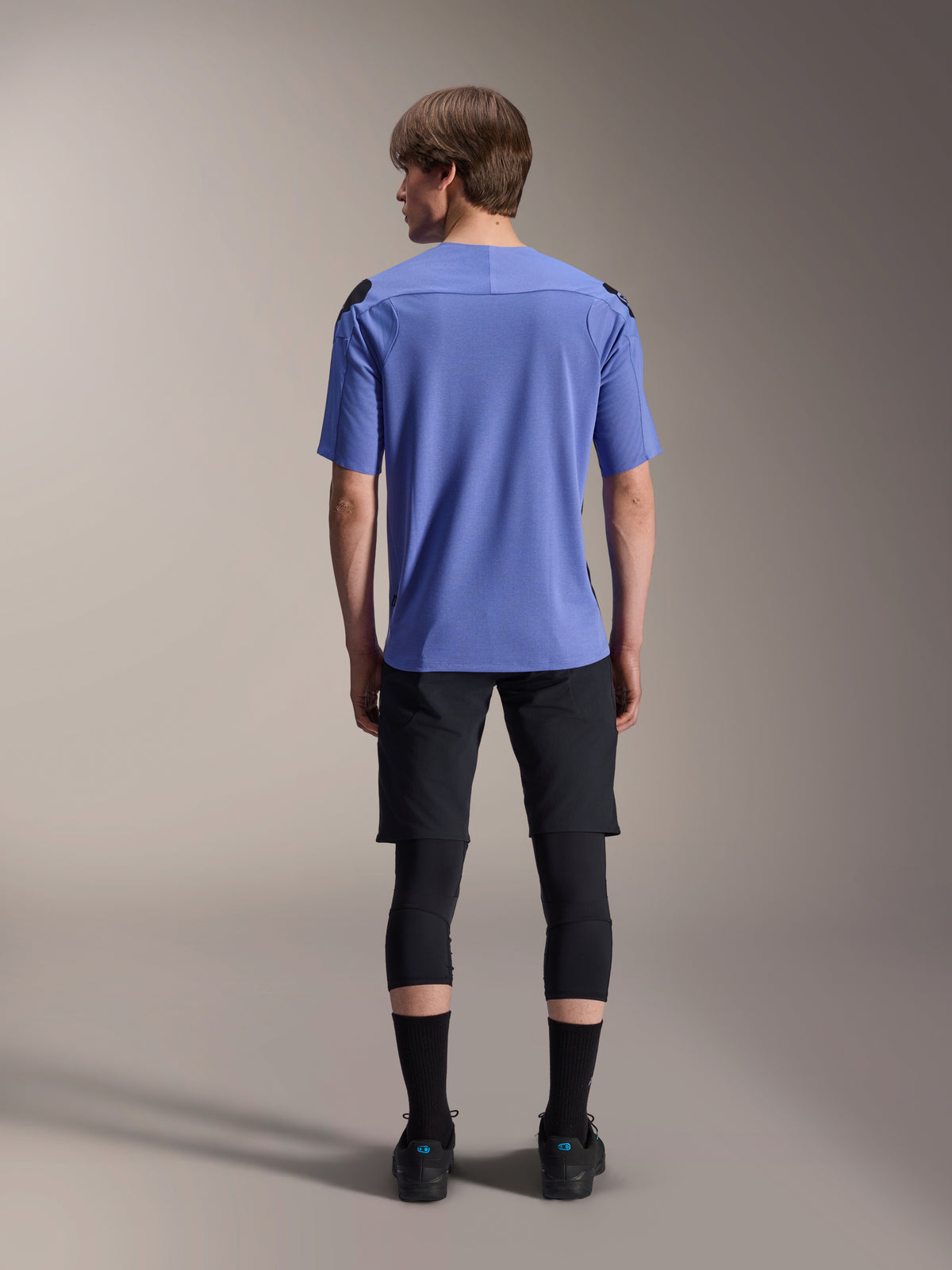 Mountain bike rider from behind wearing Alpinestars A-Aria Lite Shorts in black, paired with a blue short-sleeve jersey, black knee guards, and black socks, performance cycling gear for off-road trails