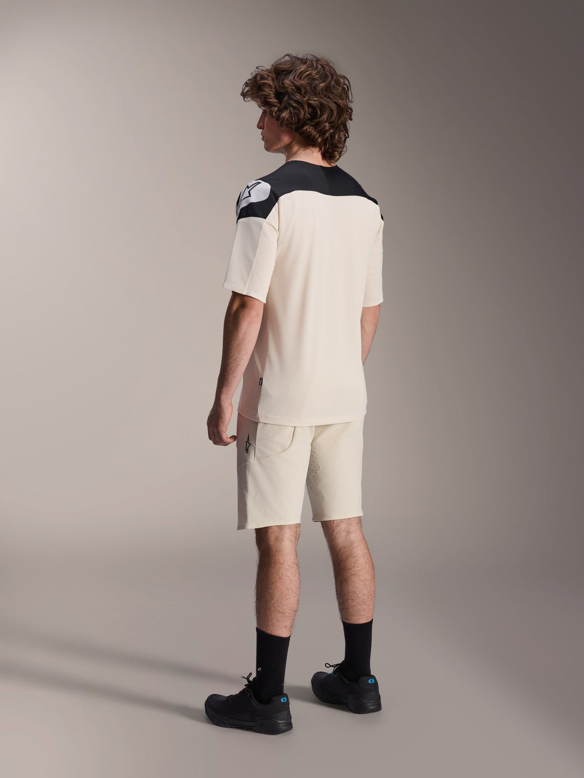 Alpinestars A-Aria Elite-shorts och matchande tröja, mountainbike-kläder, ecru med svarta detaljer, manlig modell sedd bakifrån iklädd svarta strumpor och skor, prestandakläder för terrängcykling
