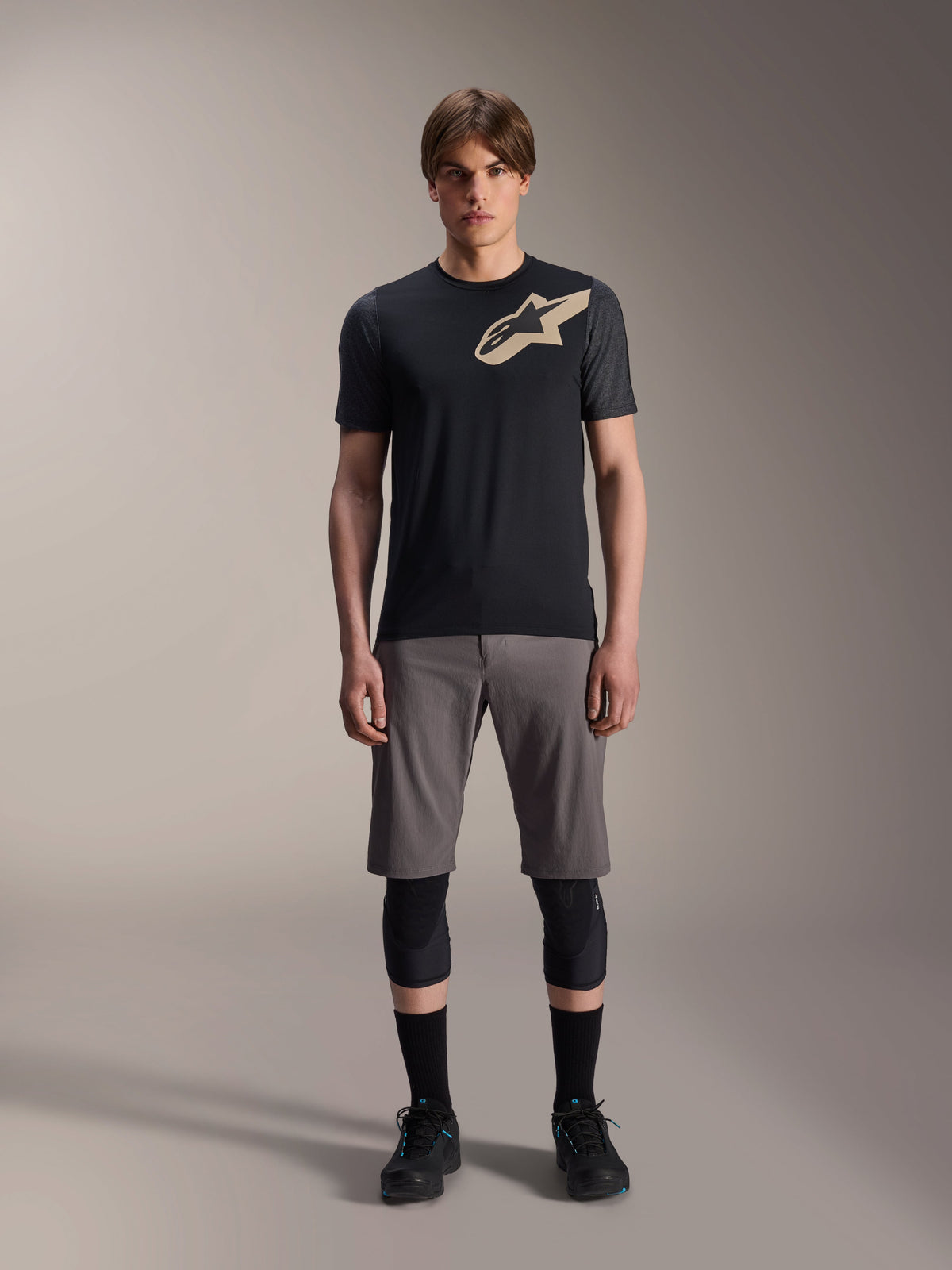Alpinestars A-Dura Lipan kortärmad tröja, svart med gråmelerade ärmar och beige Astars-logotyp på bröstet, bärs av en manlig modell med grå mountainbikeshorts och svarta knäskydd, funktionell MTB-utrustning.