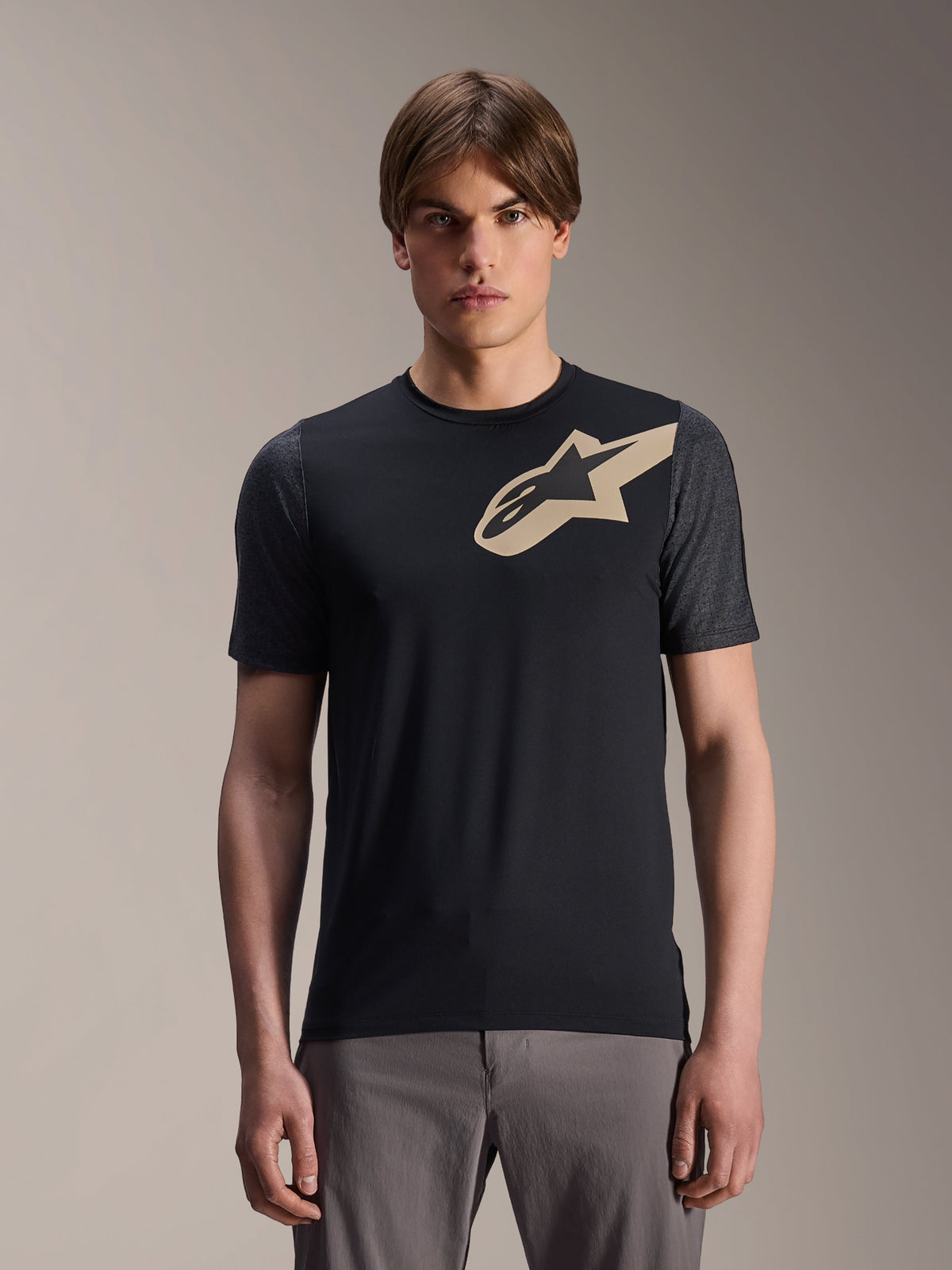 Manlig modell som bär Alpinestars A-Dura Lipan kortärmad tröja, svart MTB-tröja med beige stjärnlogotyp och mörkgrå ärmar, kombinerat med grå cykelbyxor, avslappnad funktionsbeklädnad