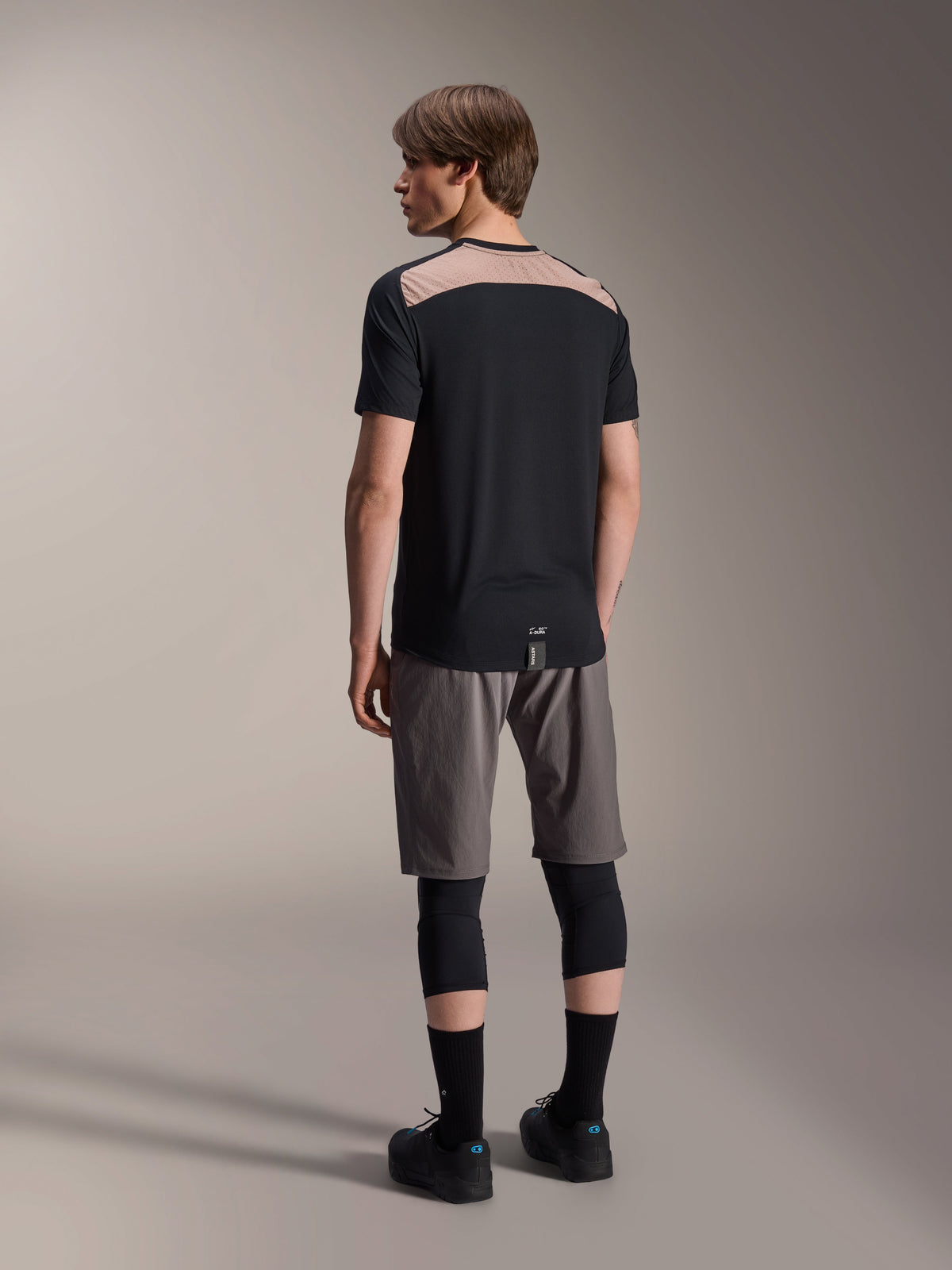 Alpinestars A-Dura Lipan kortärmad tröja i svart, ryggvy på modell som visar den ljusbruna perforerade meshpanelen över övre ryggen, kombinerad med grå mountainbikeshorts och svarta knäskydd för en komplett outfit för stigcykling.