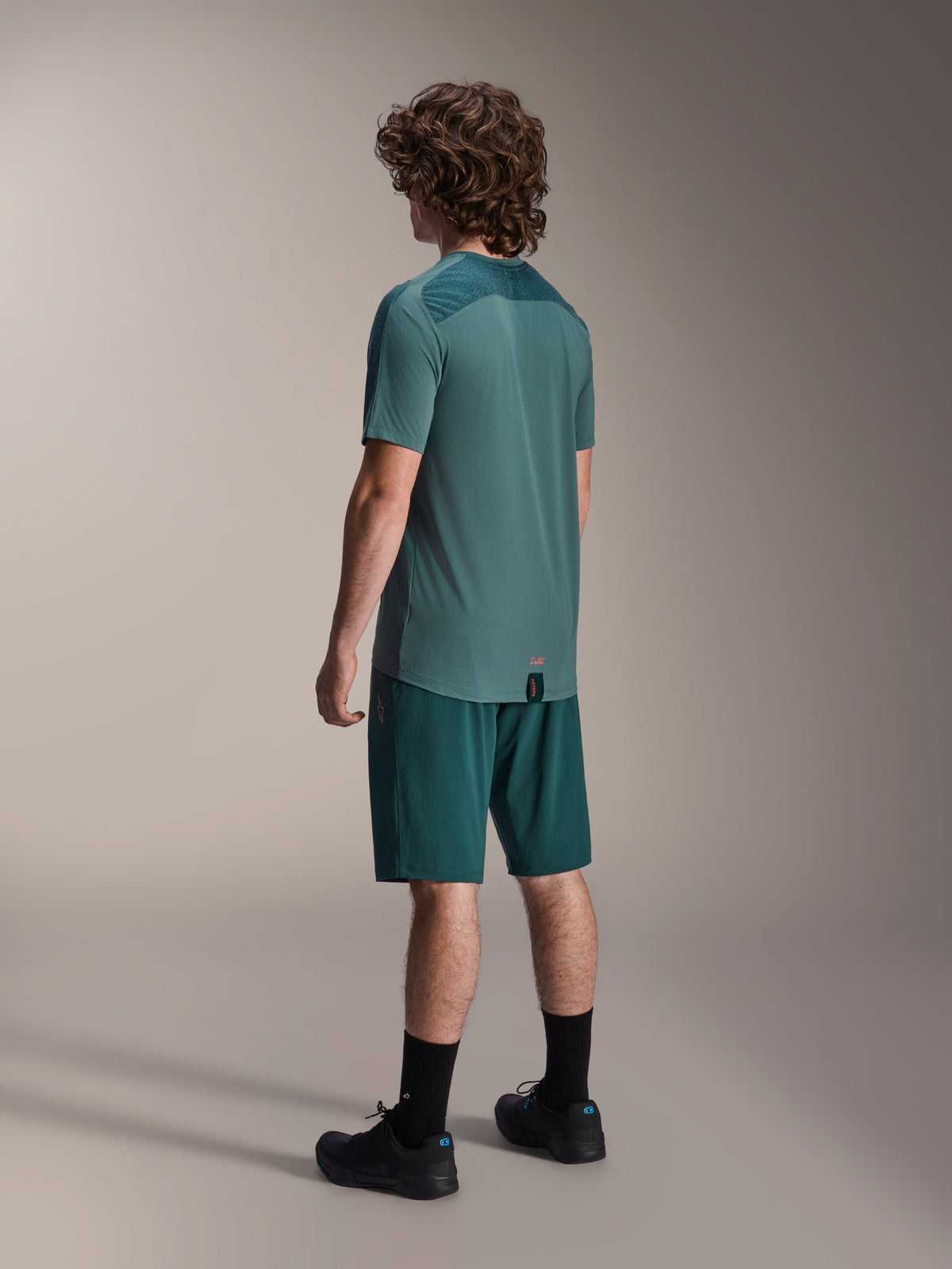 Manlig modell bär Alpinestars A-Dura Lipan kortärmad tröja i Silver Pine, sedd bakifrån, kombinerat med matchande mörkgröna mountainbikeshorts, svarta strumpor och skor, med en texturerad övre ryggpanel och ett förlängt bakstycke för extra täckning.