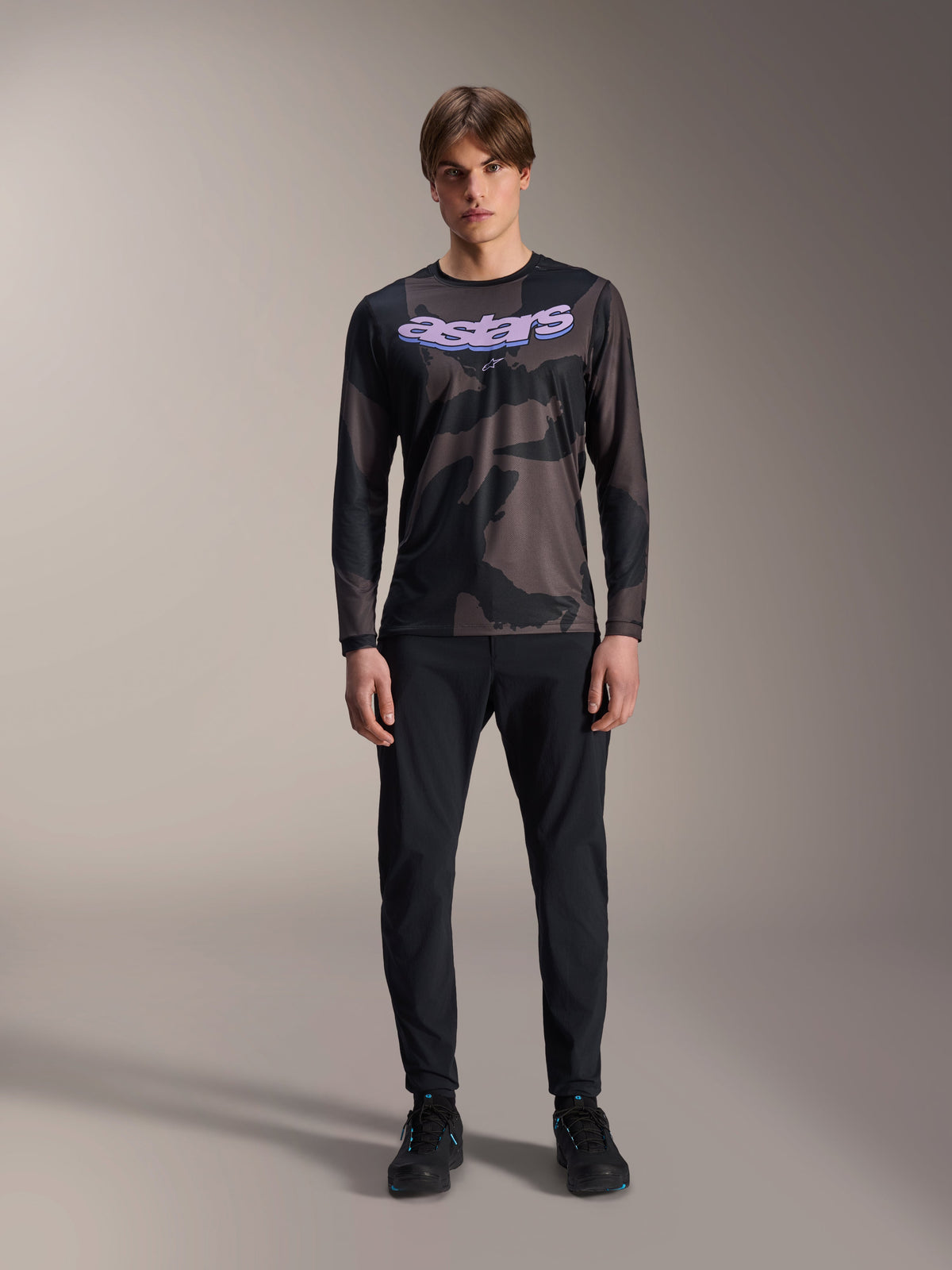 Modell klädd i Alpinestars A-Dura Hollow Camo långärmad tröja i Black Camo, med en lila 'astars'-logotyp på bröstet, matchad med svarta byxor och svarta cykelskor, mountainbike-utrustning