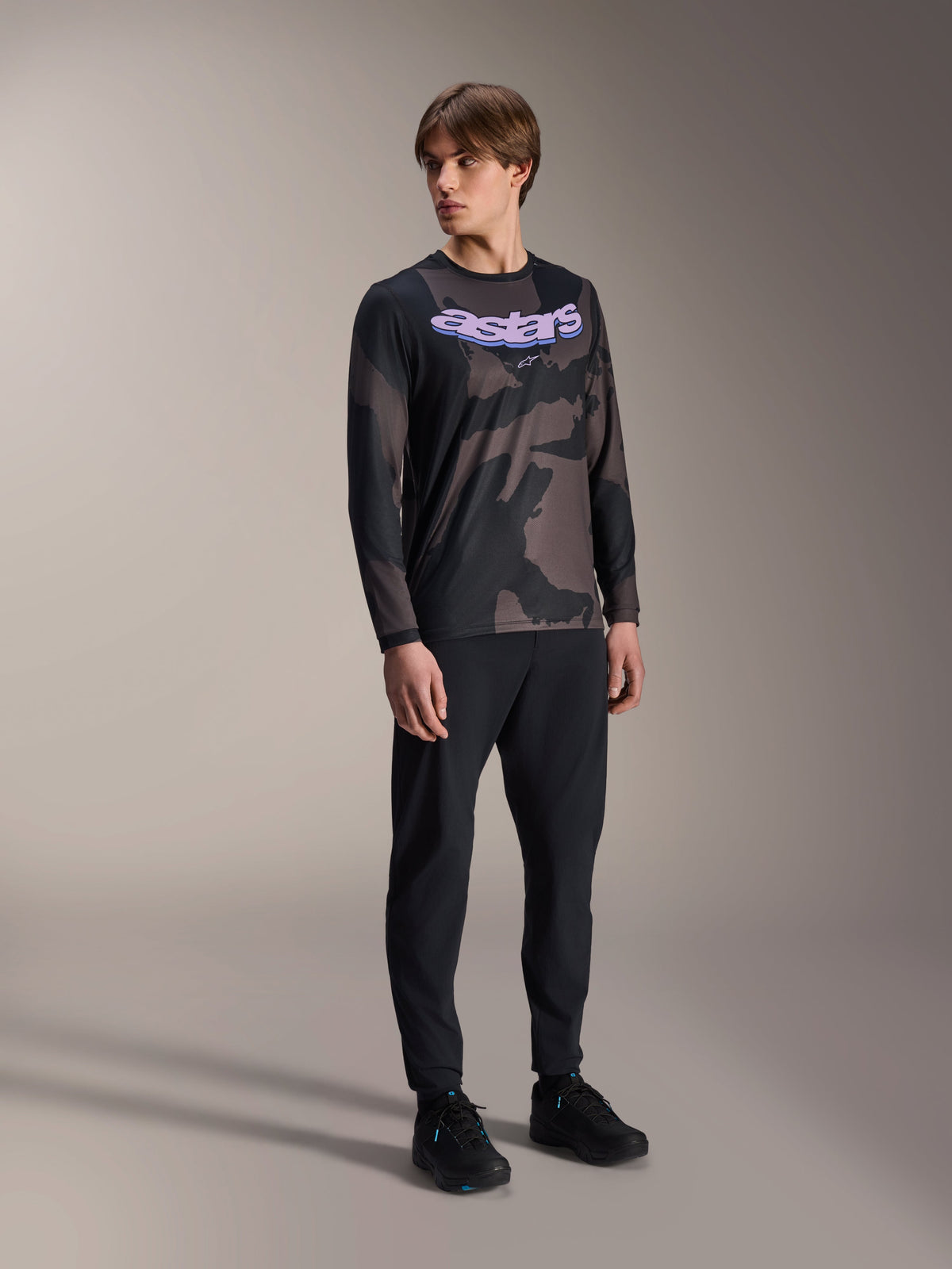 Manlig modell iklädd Alpinestars A-Dura Hollow Camo långärmad tröja, Black Camo-mönster med lila 'astars'-grafik på bröstet, kombinerat med svarta tekniska byxor och svarta skor, avslappnad MTB-utrustning