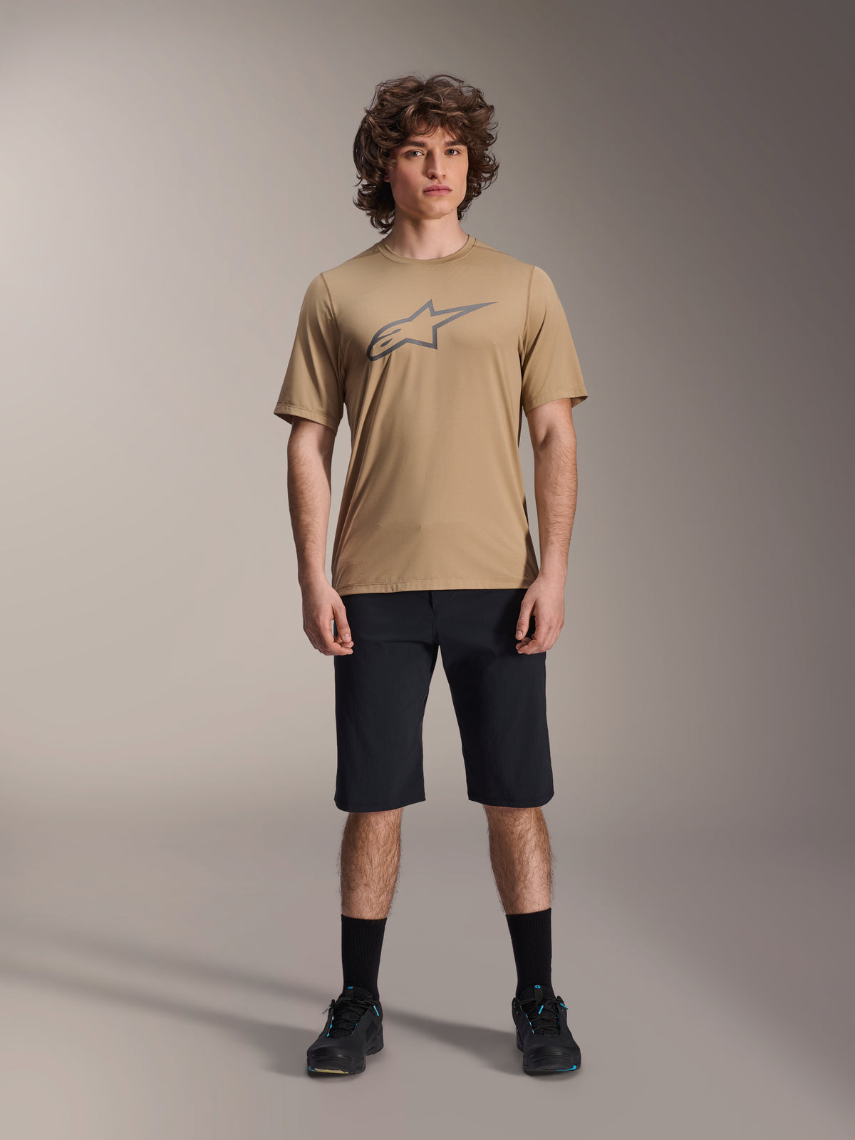 Modell som bär Alpinestars A-Dura SS Jersey i beige och svarta A-Dura Shorts, med en grå stjärnlogotyp på bröstet, matchat med svarta strumpor och mountainbikeskor, komplett MTB-utrustning i studiomiljö.