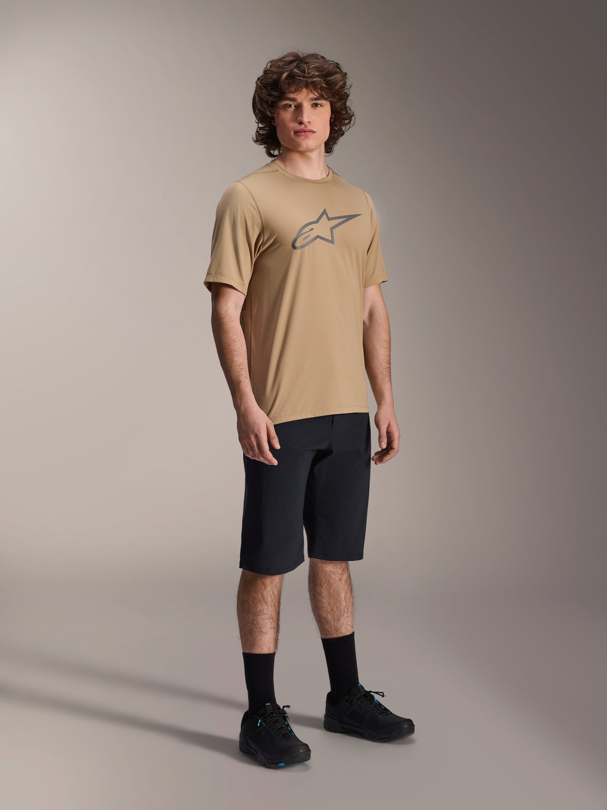 Alpinestars A-Dura Shorts, svarta mountainbikeshorts, bärs av en modell med en beige kortärmad tröja med Alpinestars logotyp, svarta strumpor och svarta skor, slitstark MTB-utrustning för terrängkörning