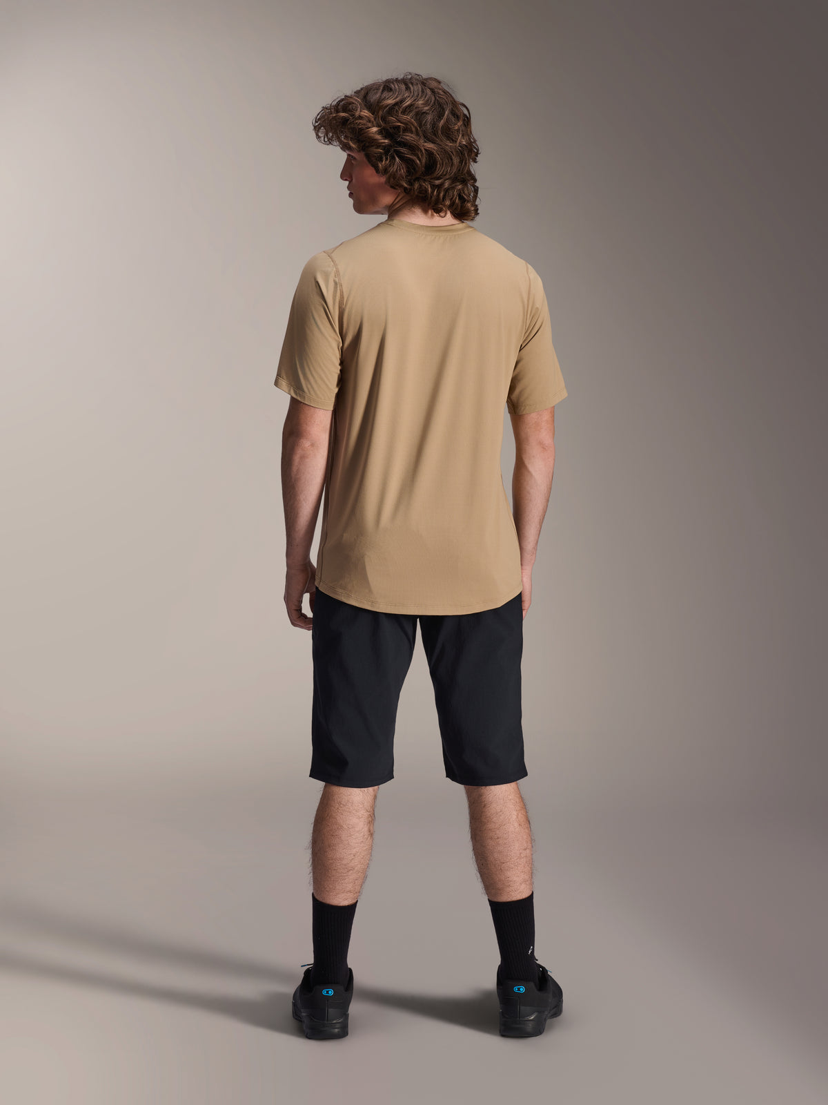 Manlig modell sedd bakifrån bärande Alpinestars A-Dura Shorts i svart och en sandfärgad A-Dura kortärmad tröja, kombinerat med svarta strumpor och mountainbikeskor, funktionell MTB-outfit för trail