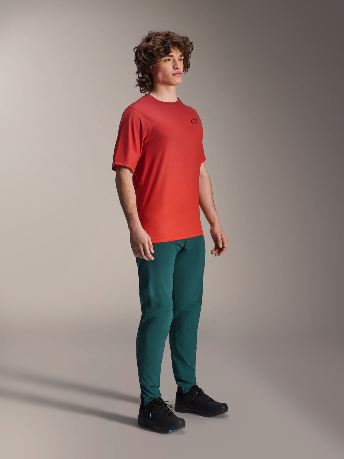 Modell som bär Alpinestars A-Dura-byxor i Dark Pine, kombinerat med en röd A-Dura Rise kortärmad tröja och svarta MTB-skor, atletiska mountainbike-kläder i studiomiljö