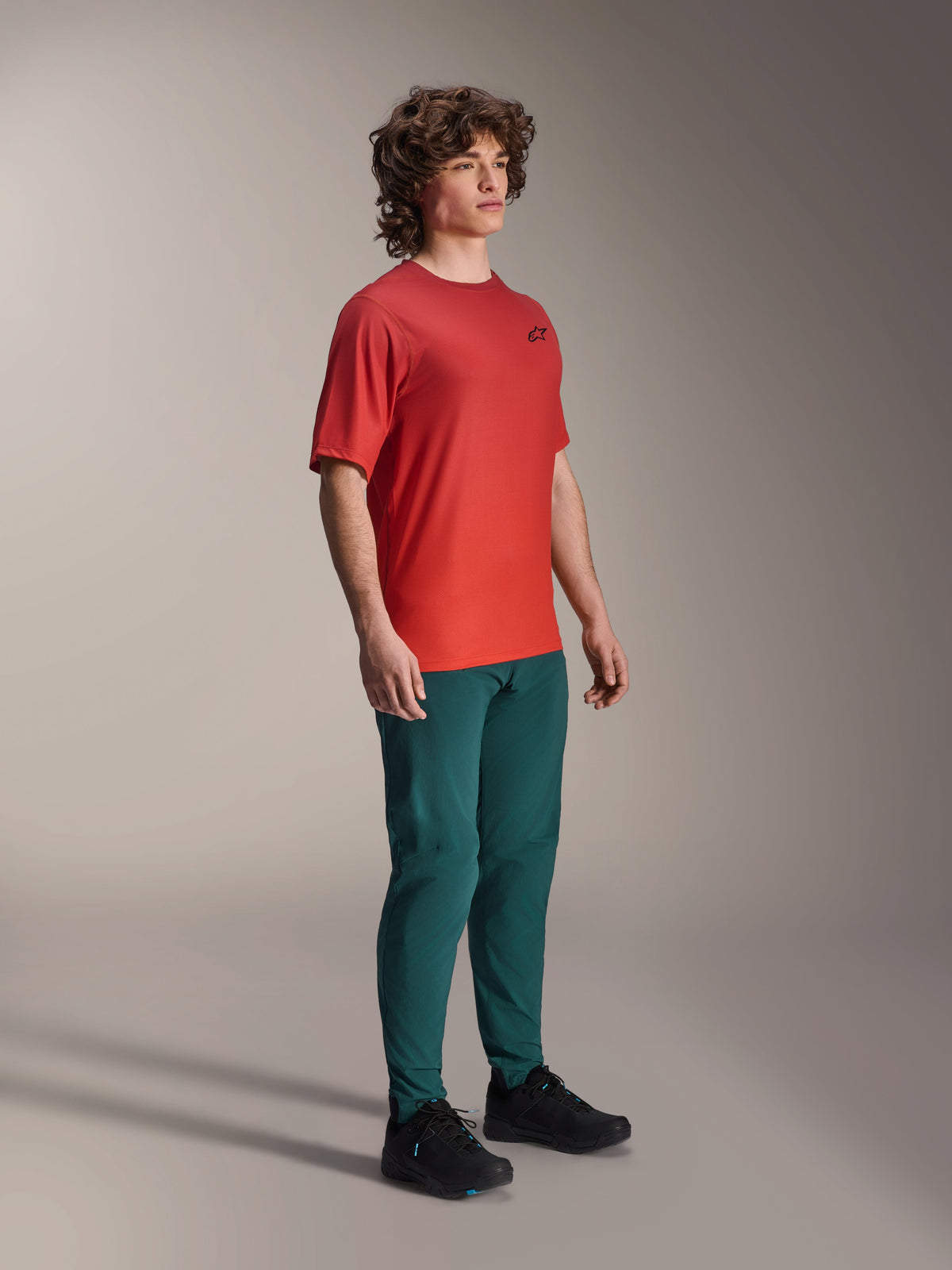 Alpinestars A-Dura Rise Jersey, kortärmad, Spicy Orange, med en liten svart logotyp på bröstet, buren med mörka blågröna byxor och svarta skor, lätta fukttransporterande mountainbike-kläder