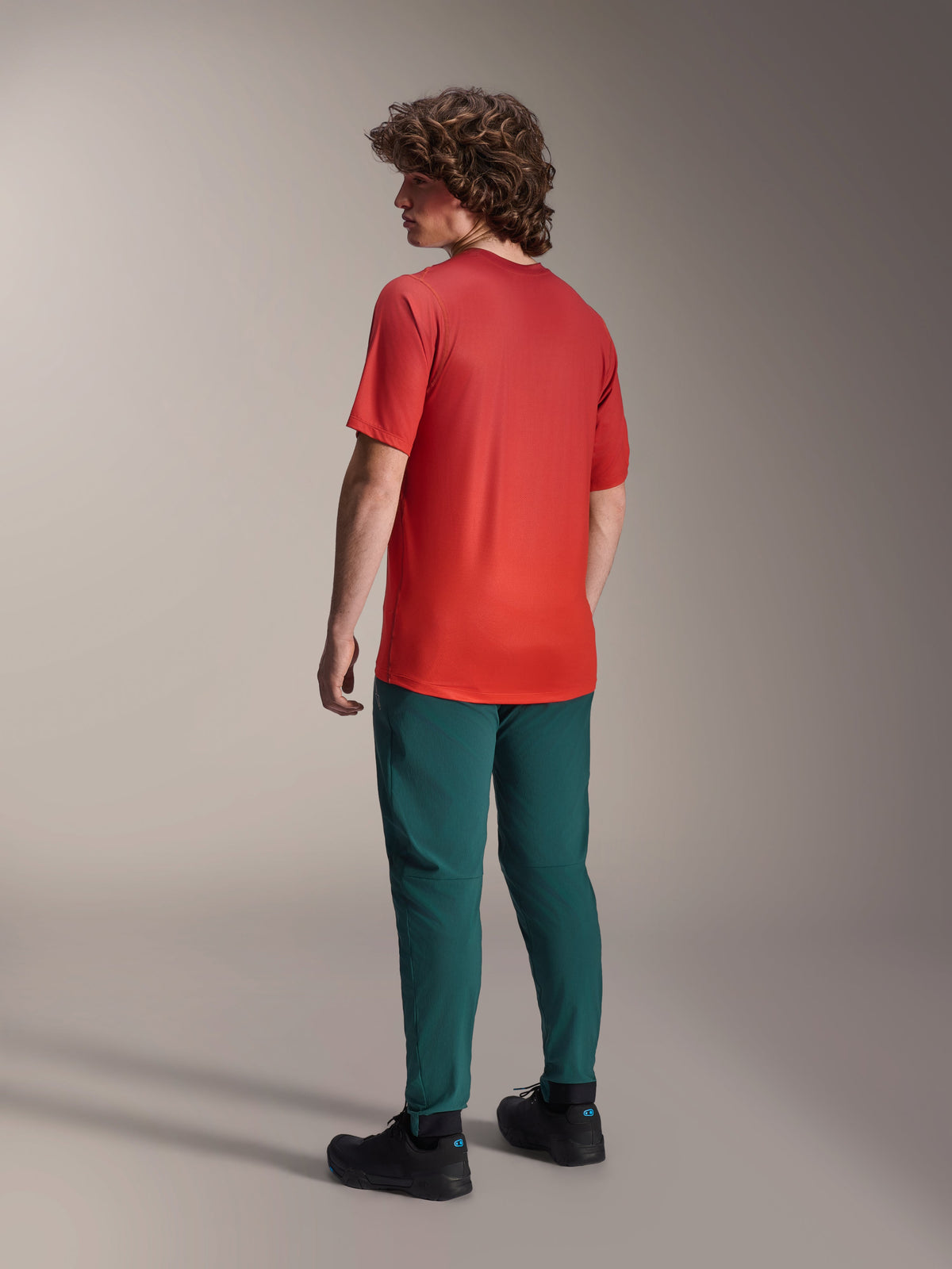 Alpinestars A-Dura Rise Tröja - Kortärmad i Spicy Orange, ryggvy av modell som bär cykeltröjan med mörkgröna mountainbikebyxor och svarta skor, med förlängt ryggslut och prestandapassform för terrängcykling
