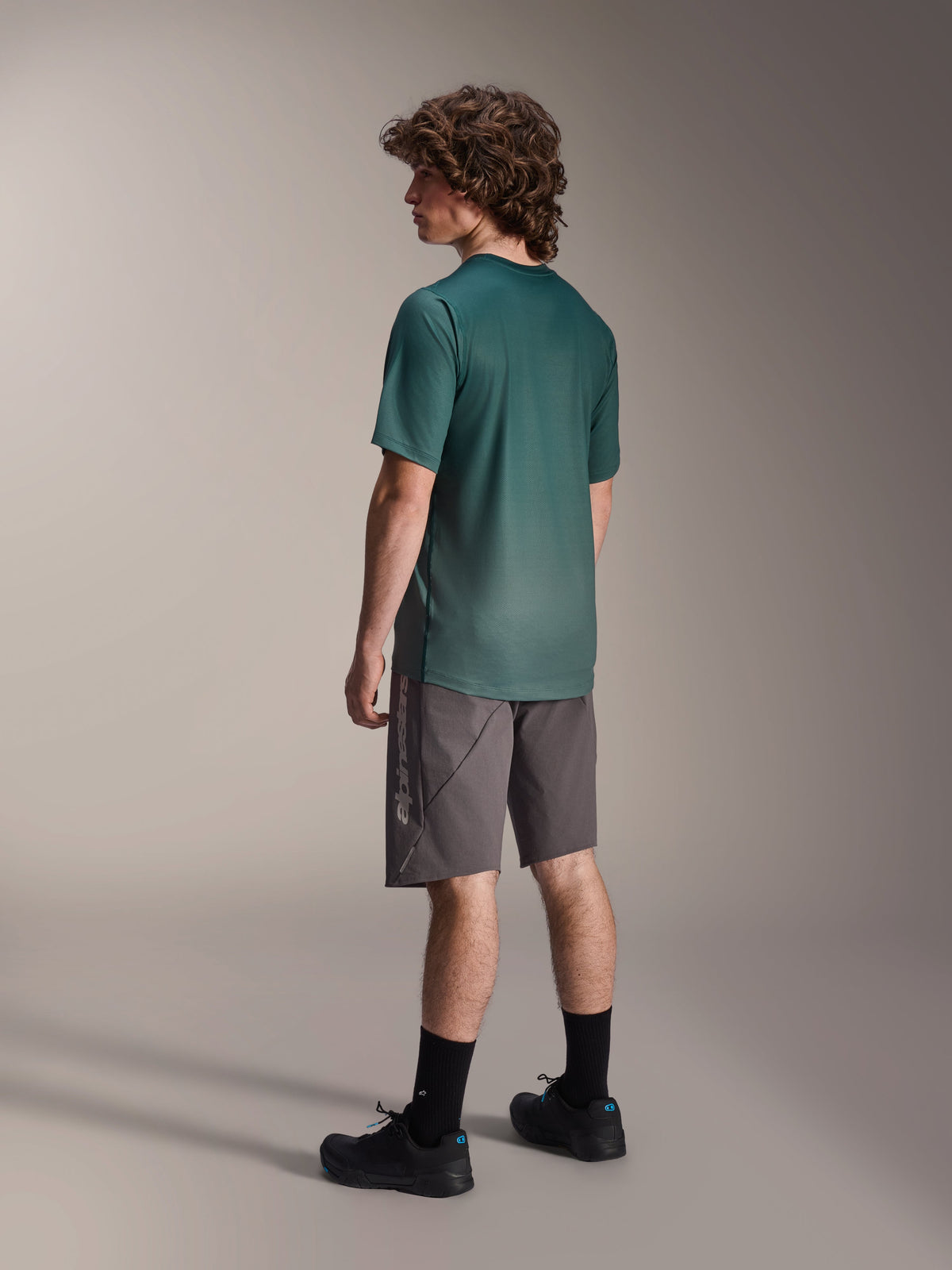 Alpinestars A-Dura Rise tröja, kortärmad MTB-tröja, Silver Pine-grön, bärs med grå shorts och svarta skor, vy bakifrån som visar förlängt bakstycke och normal passform, lätt fukttransporterande material för stigcykling