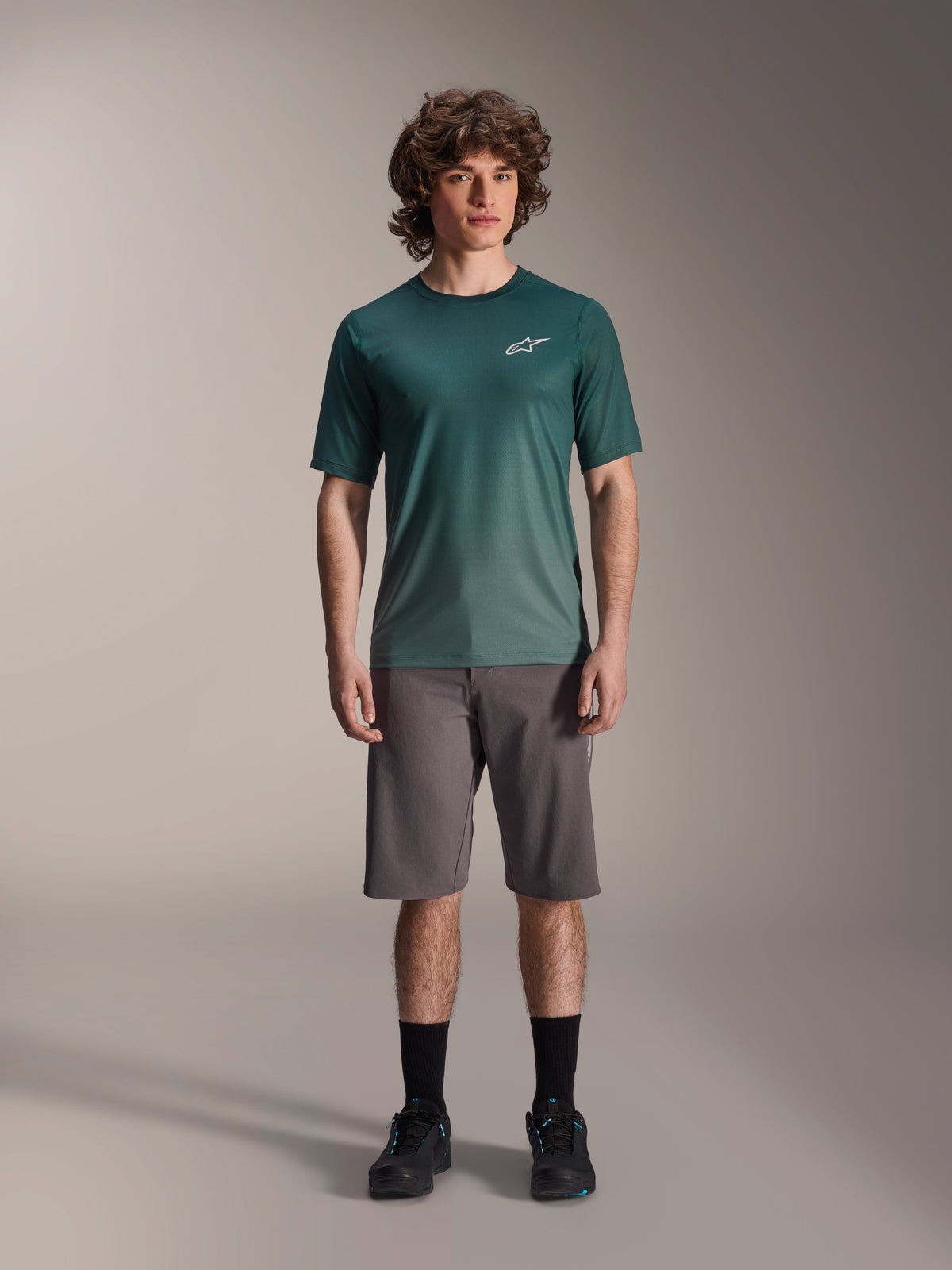 Manlig modell i Alpinestars A-Dura Rise kortärmad tröja i Silver Pine, grön cykeltröja med liten vit logotyp på bröstet, matchad med grå mountainbikeshorts och svarta skor, avslappnad MTB-utrustning