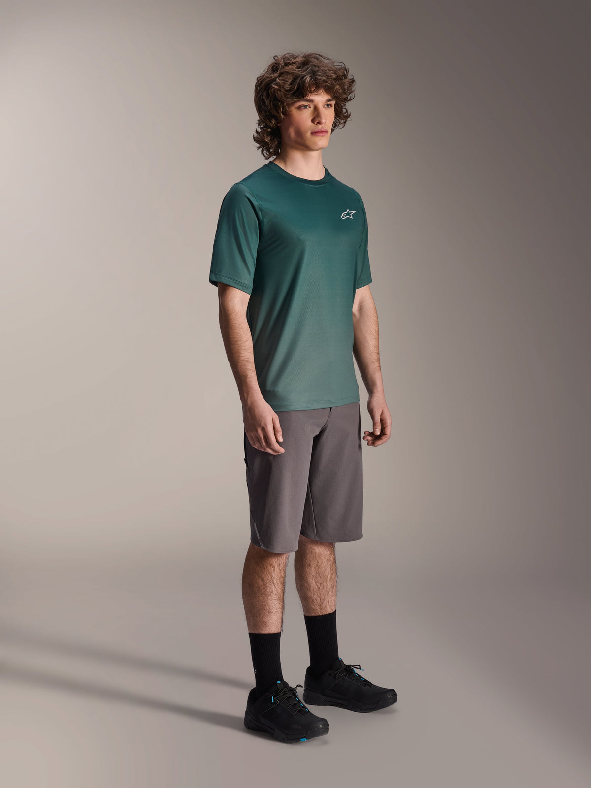 Alpinestars A-Dura Rise Jersey - Short Sleeve i Silver Pine, grön mountainbiketröja bärs av en modell med gråa shorts och svarta sneakers, avslappnad MTB-utrustning för stigcykling med en liten vit logotyp på bröstet
