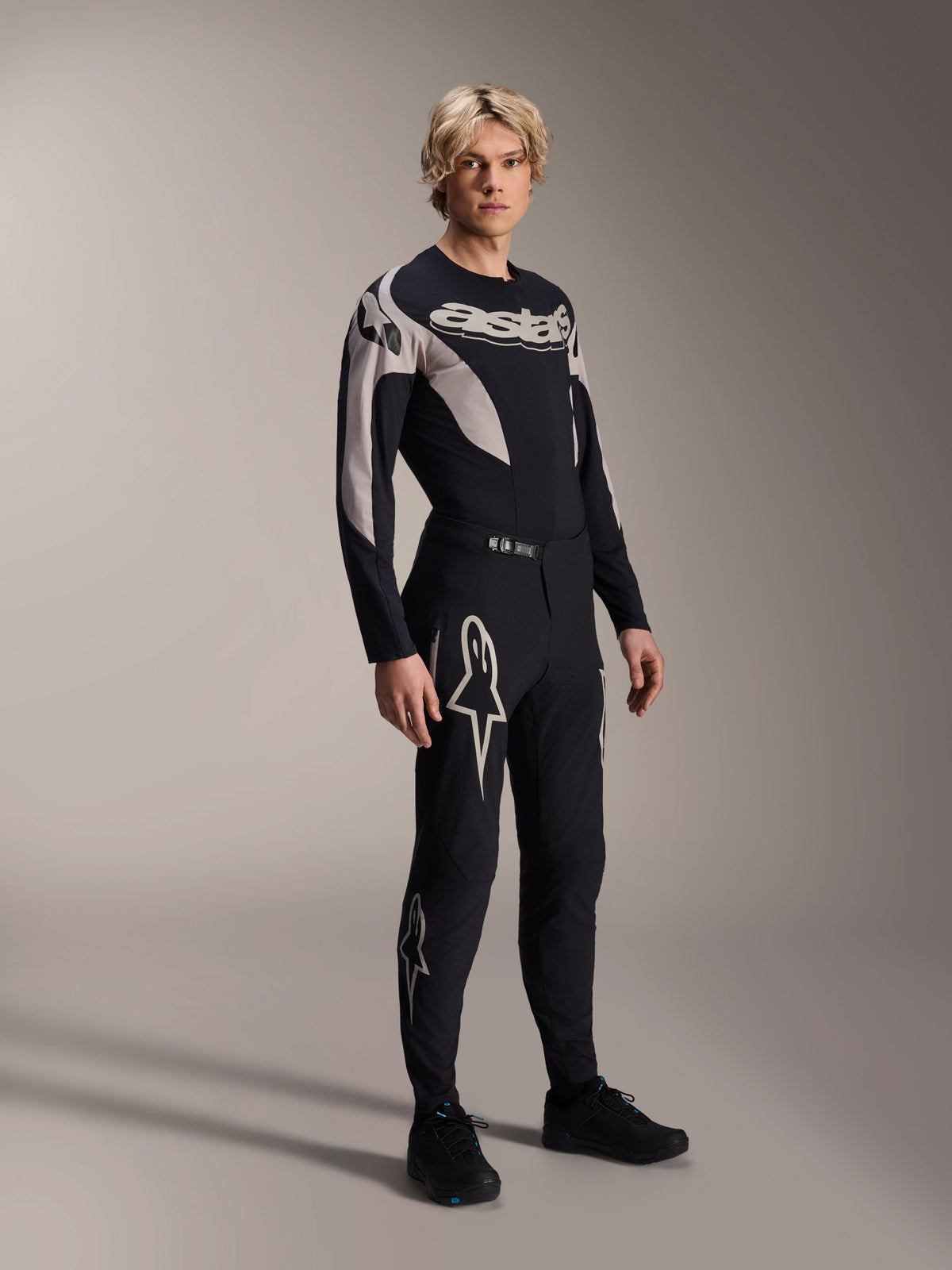 Manlig modell iklädd Alpinestars A-Supra Melt-byxor och matchande långärmad tröja, svart med ljusgrå kurvade mönster, framträdande vita Astars-logotyper på benen och ordmärke på bröstet, professionell MTB-racingoutfit.