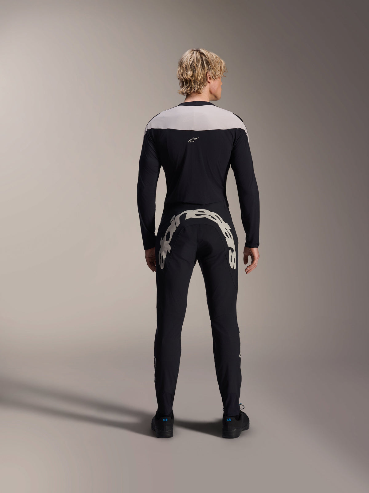 Alpinestars A-Supra Melt-byxor, svarta, baksida som visar det stora vita Alpinestars-ordmärket över bakoket, kombinerat med en matchande långärmad tröja och svarta cykelskor, högpresterande mountainbike-utrustning för terrängkörning.