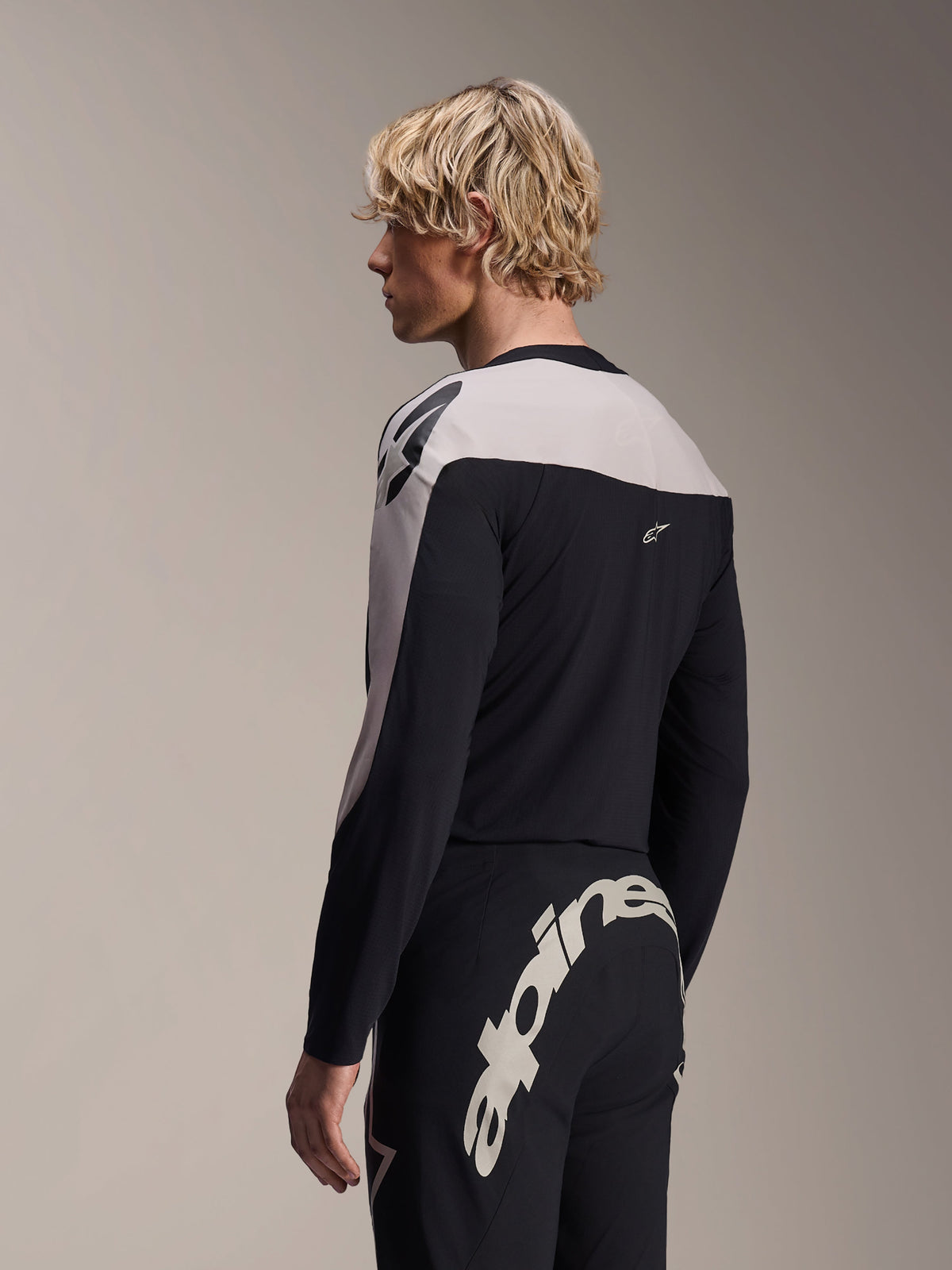 Manlig modell bär Alpinestars A-Supra Melt långärmad tröja och A-Supra Melt-byxor, svart och ljusgrått MTB-racingkit, med ett stort vitt Alpinestars-ordmärke över byxornas bakok