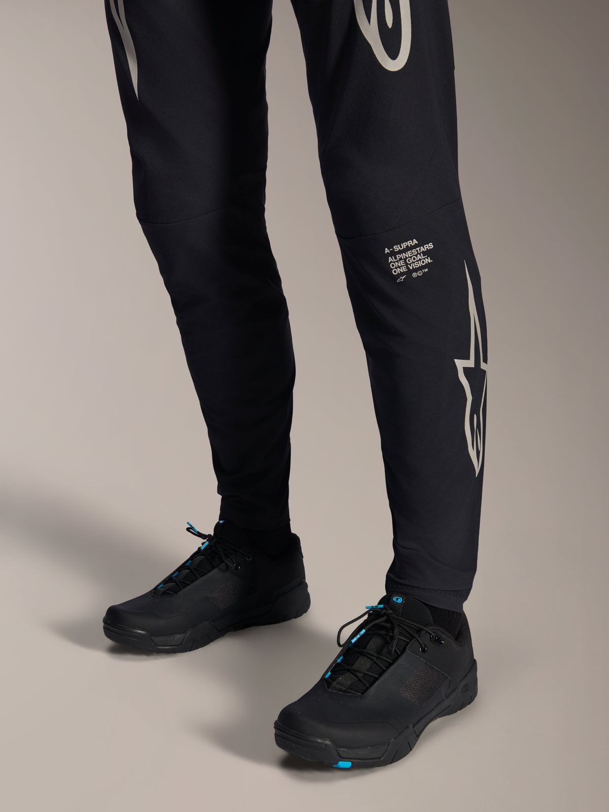 Alpinestars A-Supra Melt-byxor, svarta mountainbikebyxor, med vitt Alpinestars-ordmärke och logotyp på benet, bärs med svarta cykelskor med blå detaljer, högpresterande terrängutrustning