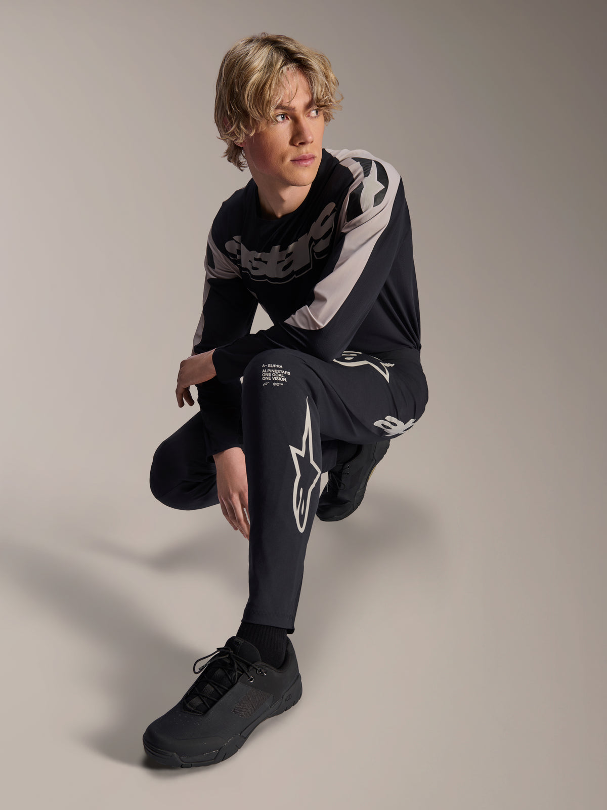 Alpinestars A-Supra Melt-byxor och långärmad tröja, svarta och grå, med framträdande vit branding och stjärnlogotyper, buren av en manlig modell i en hukande pose, matchande MTB-racingutrustning