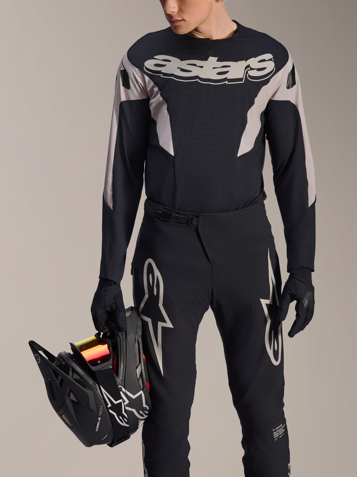 Alpinestars A-Supra Melt MTB-racingkit, långärmad tröja och byxor, svarta och grå med vita Astars-logotyper, modell som håller en svart fullfacehjälm och bär matchande handskar, högpresterande utrustning för downhill- och enduro-mountainbike