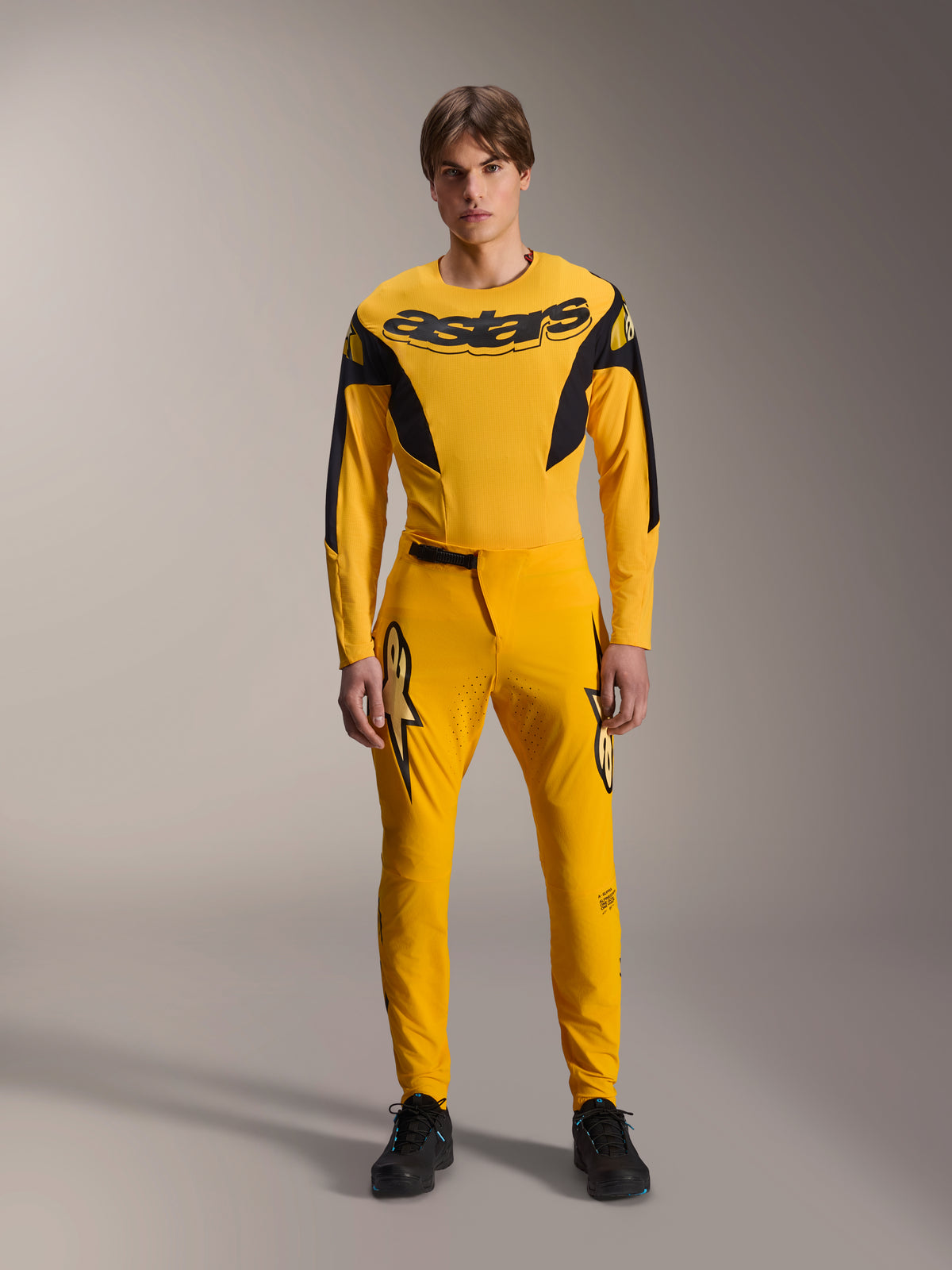 Limited Edition A-Supra Melt Gold-utrustningsset, Alpinestars mountainbike-kläder, mörkgul och svart, modell som bär långärmad tröja med tydlig ”astars”-logotyp på bröstet och matchande funktionsbyxor med laserskuren ventilation på insidan av låren och en säker ratch-stängning i midjan, professionellt racingkit för MTB