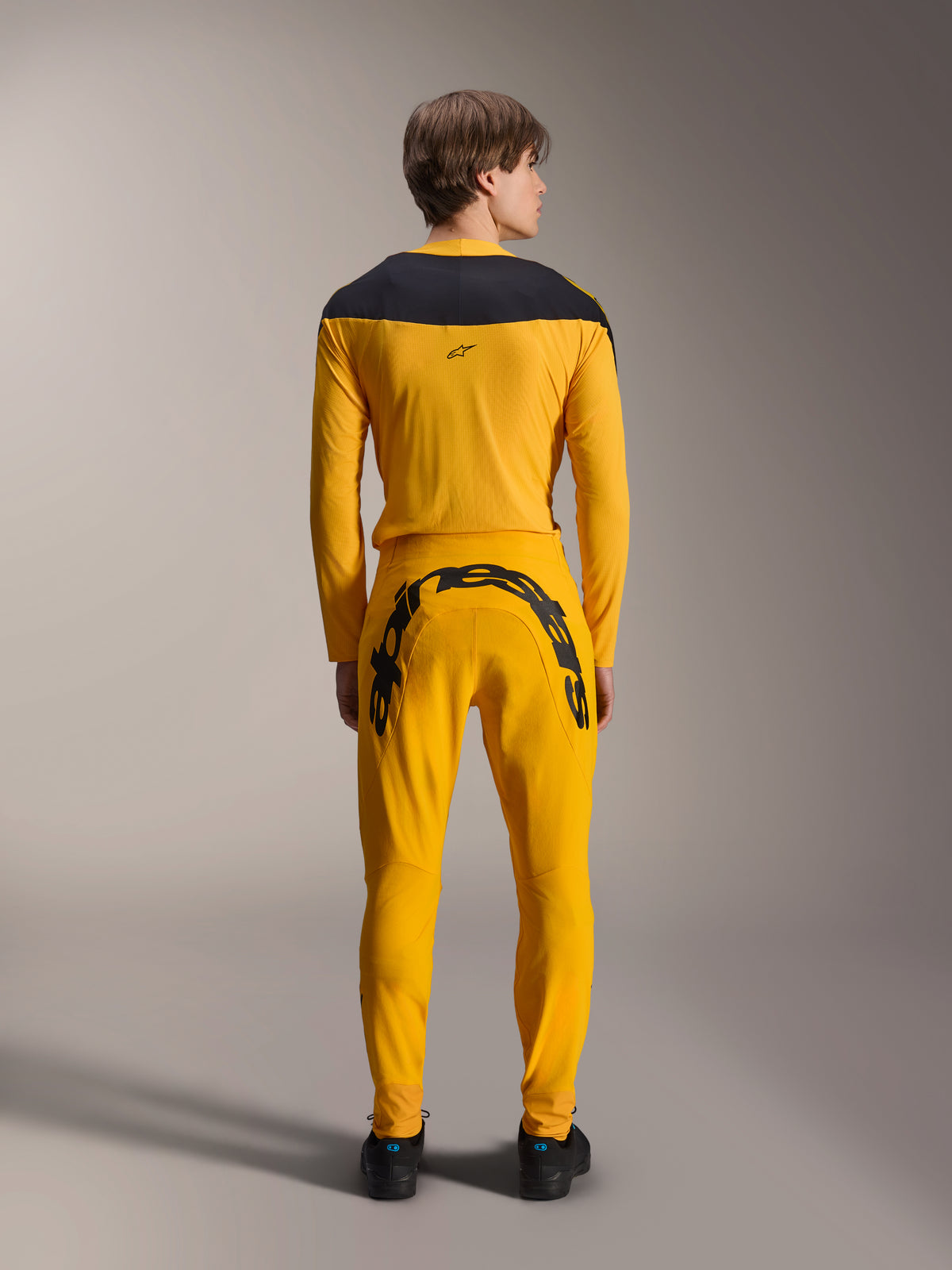 Alpinestars Limited Edition A-Supra Melt Gold Gear-Set, vy bakifrån, mörkgul långärmad tröja med svart axelok, matchande mörkgula byxor med ett stort svart Alpinestars-ordmärke över bakoket, svarta skor