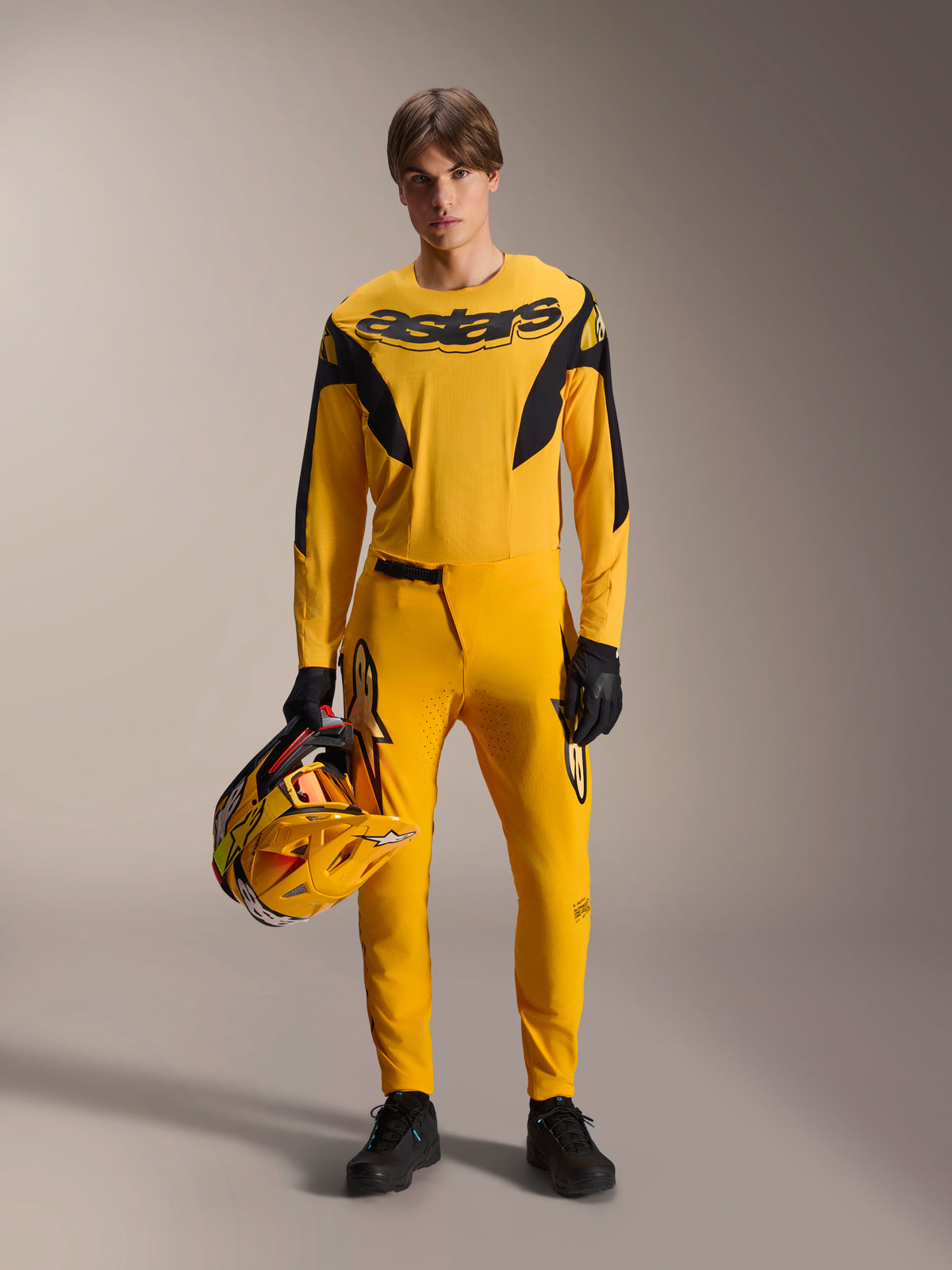 Modell som bär Alpinestars Limited Edition A-Supra Melt Gold MTB-kit, gul långärmad tröja med svart 'astars'-logotyp, matchande gula byxor med svarta detaljer, håller en gul och svart racinghjälm, svarta handskar och skor, högpresterande utrustning för mountainbike