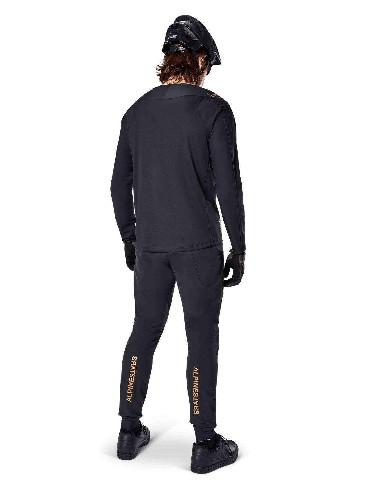 Bakifrån-vy av en mountainbikecyklist som bär Alpinestars A-Aria Polartec Switch långärmad tröja i svart, matchad med svarta MTB-byxor med beige Alpinestars-logotyper på vaderna, svart hjälm och handskar, professionell cykelutrustning för terrängkörning