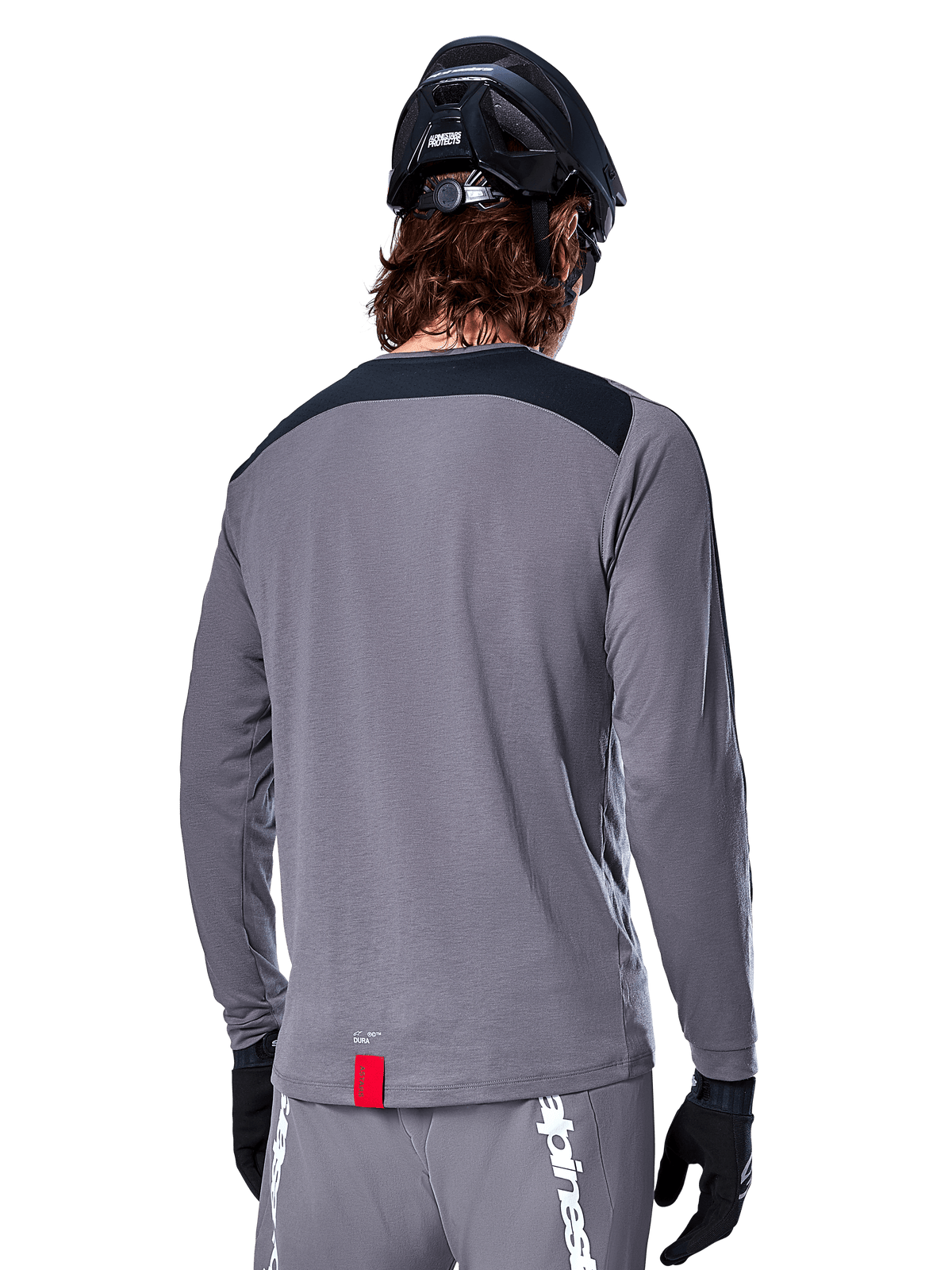 Alpinestars A-Dura Dri Astar långärmad tröja, mörkgrå MTB-tröja sedd bakifrån, med en svart axelpanel och liten röd märkesetikett, buren med en svart mountainbikehjälm och handskar