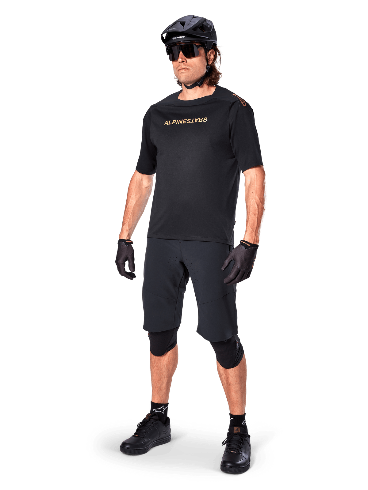 Alpinestars A-Aria Polartec Switch kortärmad tröja, svart, med speglad guldlogotyp på bröstet, bärs av en cyklist med svarta cykelshorts, hjälm och knäskydd, teknisk MTB-utrustning för terrängprestanda